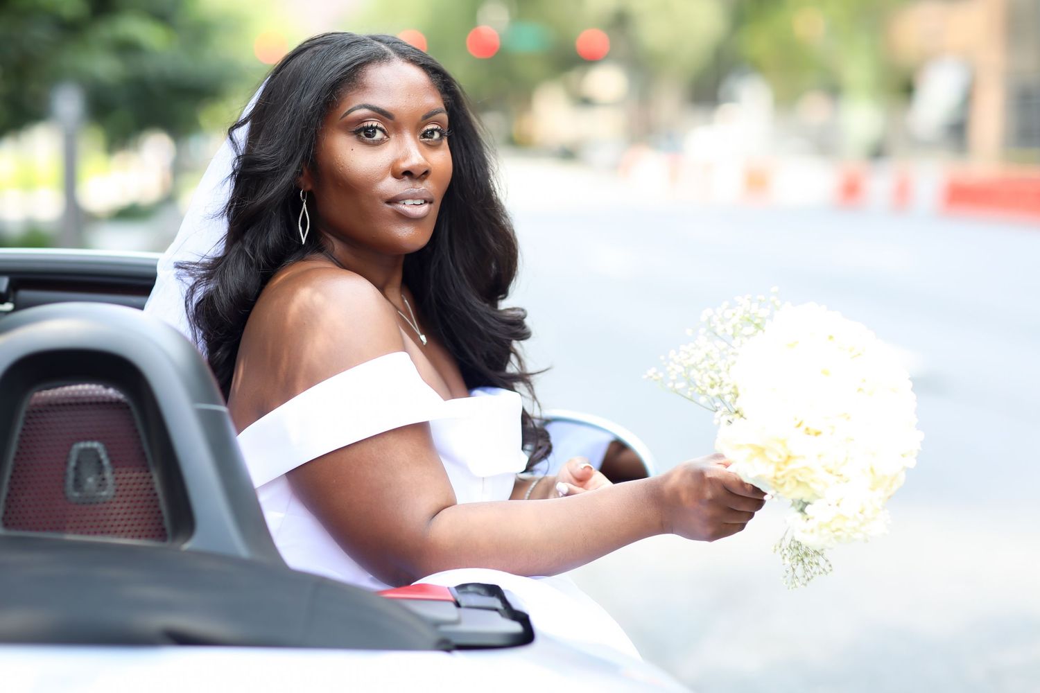 Bride in white dress waves from convertible car during wedding celebration.