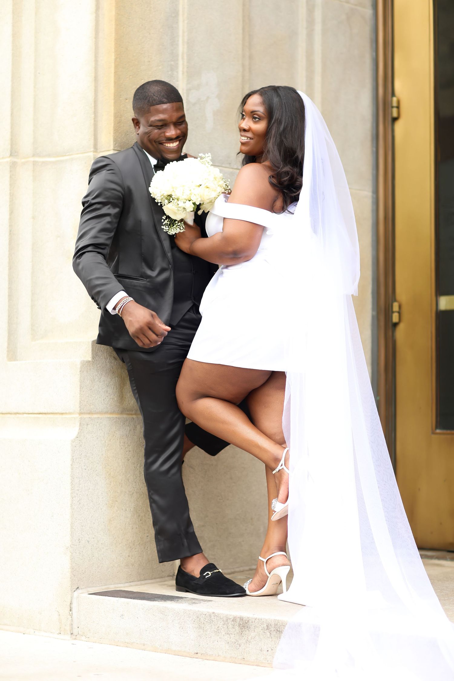 Playful wedding couple poses against stone building columns in formal attire.