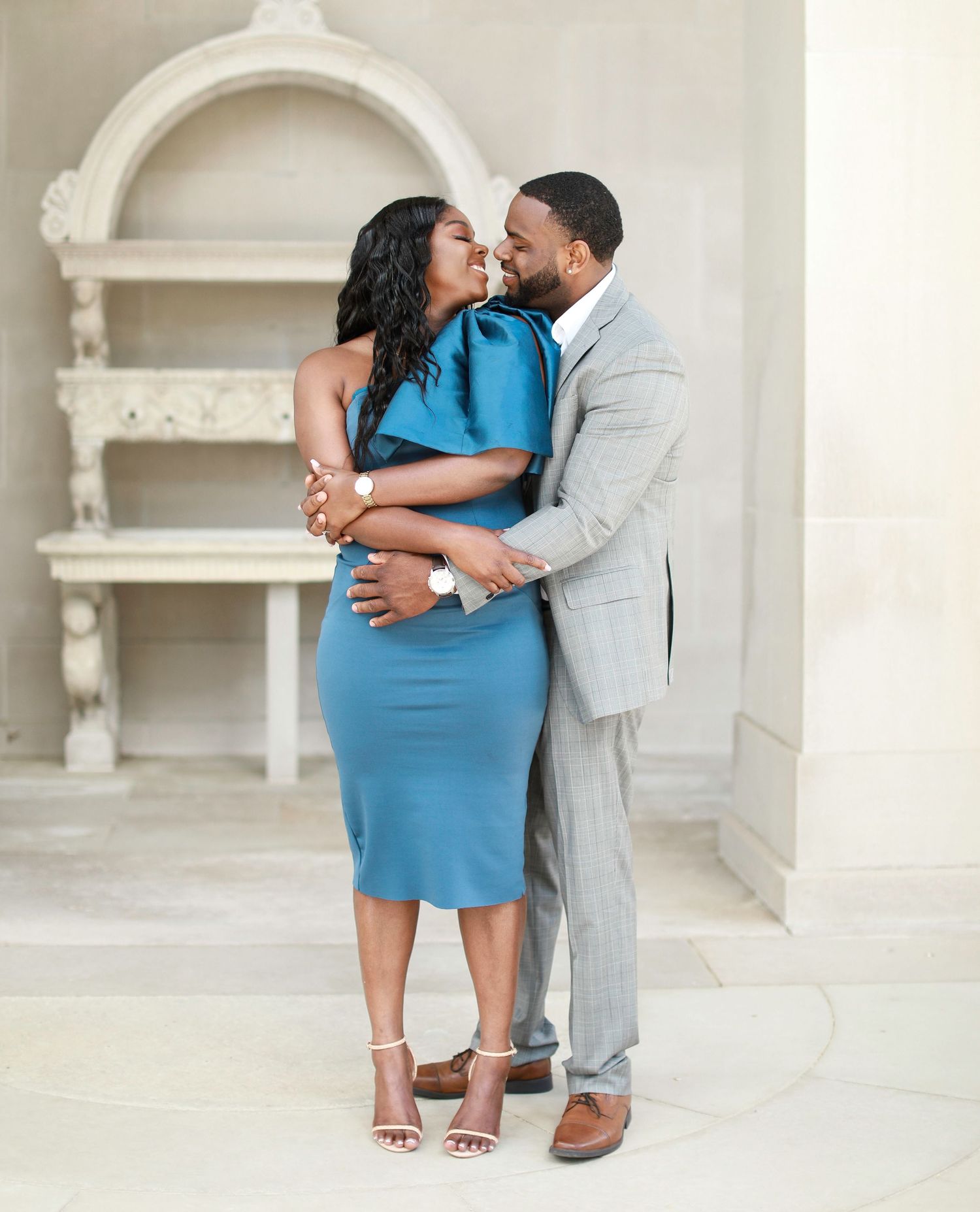 Couple in blue dress and grey suit posing together against white architectural backdrop.