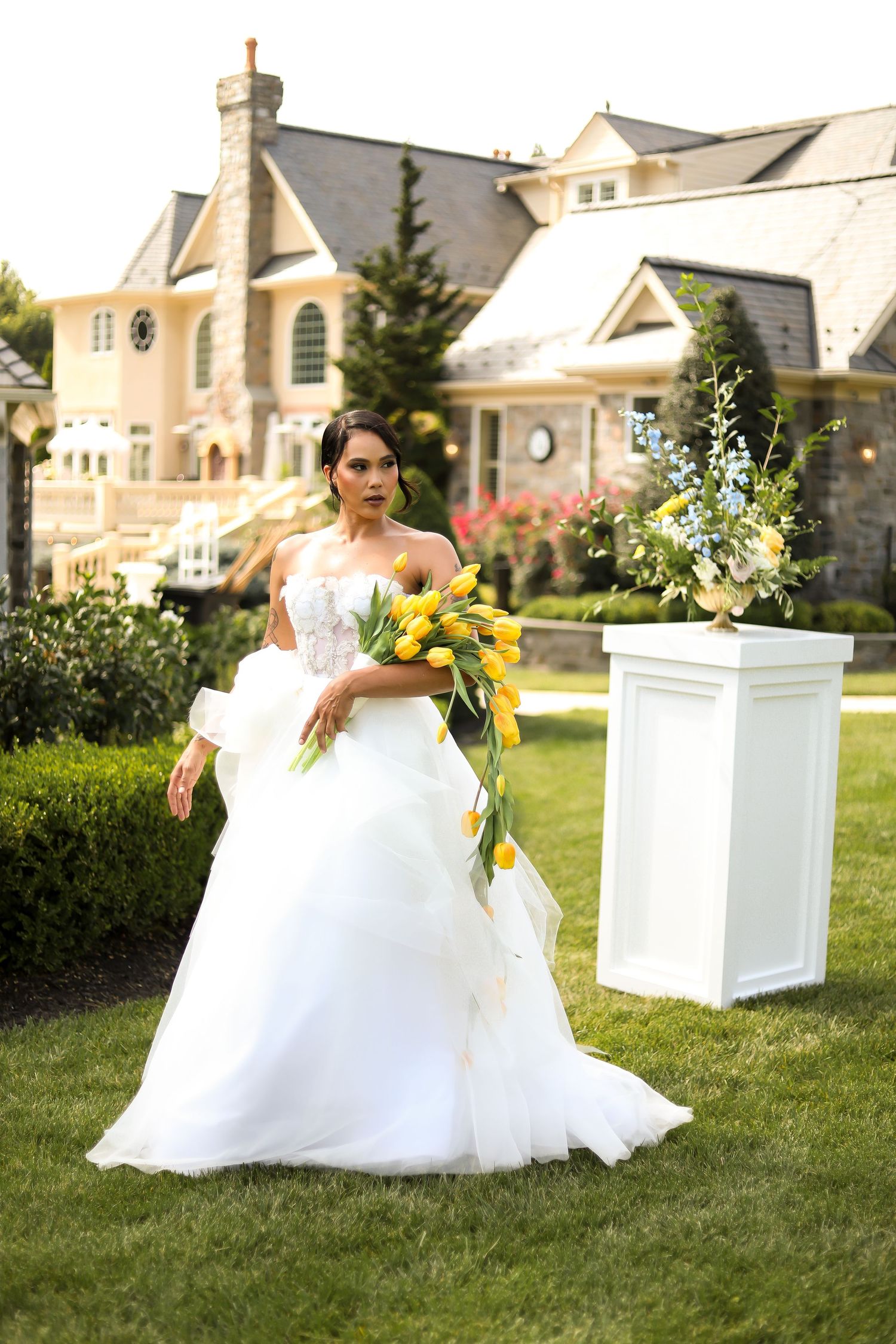 A bride in a flowing white dress poses with yellow flowers near decorative white pillars.