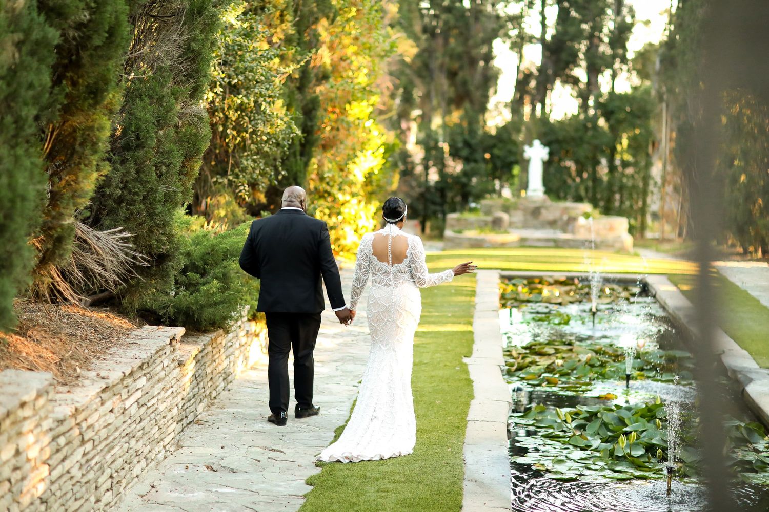Wedding couple walking through a scenic garden path with golden sunlight filtering through trees.