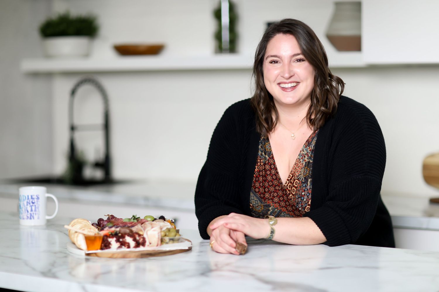 A person in a black top smiles while sitting at a bright white kitchen countertop with plates of food.