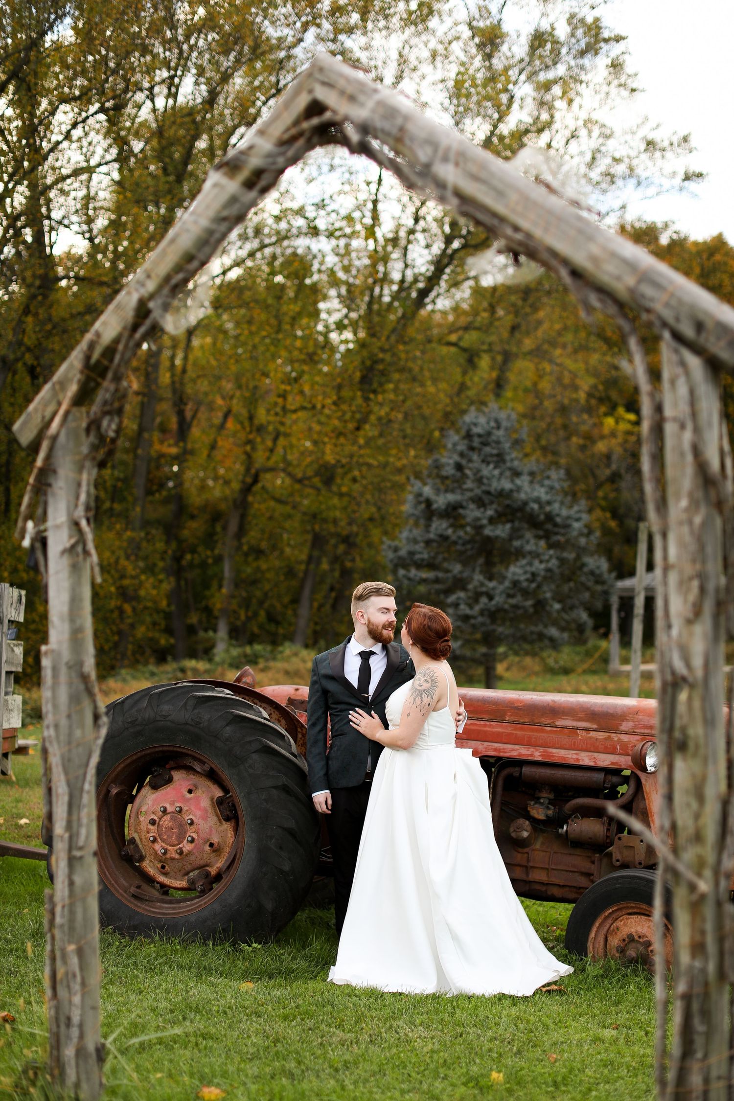 A wedding couple poses together in a rustic outdoor setting with an old tractor and wooden archway.