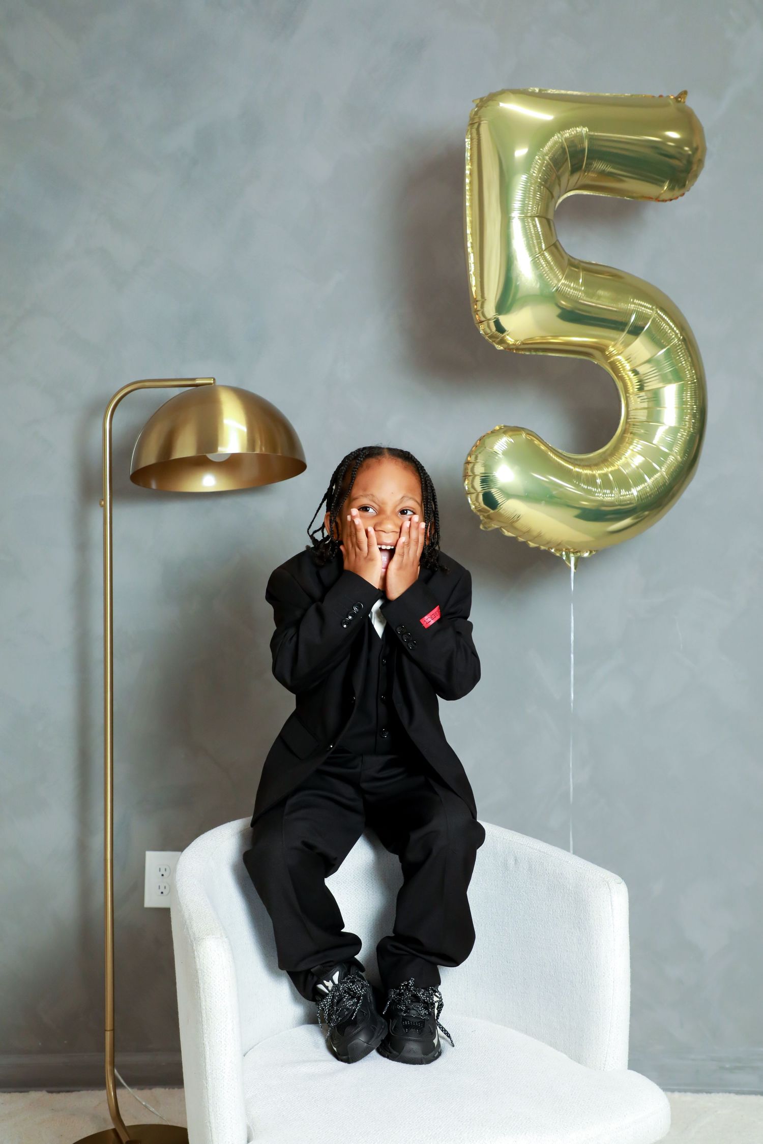 A child sits in a white armchair next to a gold floor lamp and a large gold number 5 balloon against a gray wall.