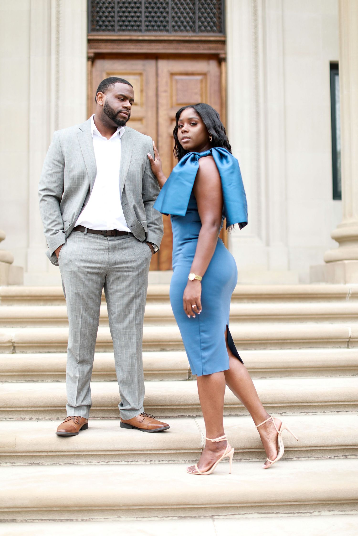 Couple in formal attire posing on elegant stone steps with grey suit and blue dress.