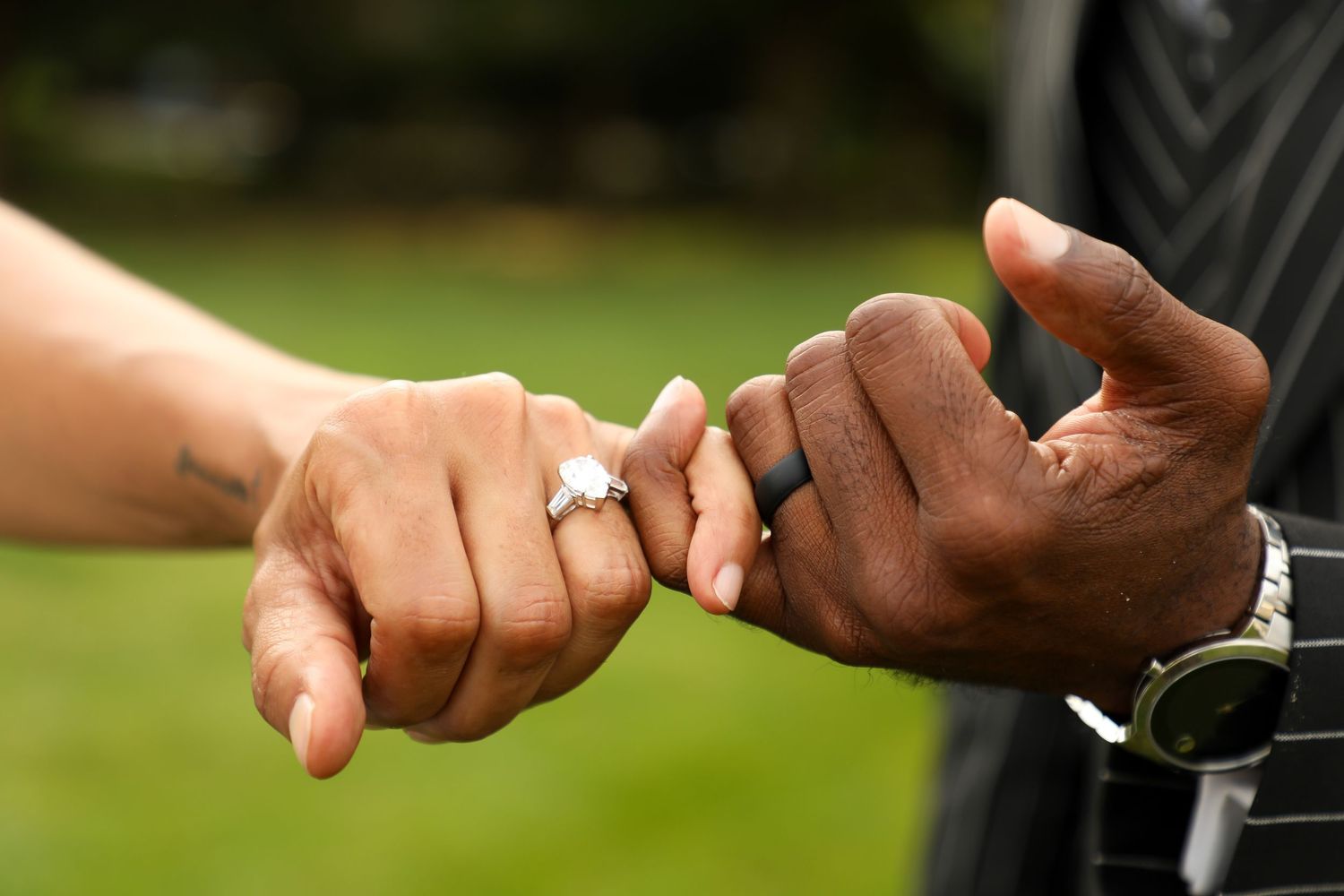 Close up of intertwined hands wearing rings against green outdoor background.