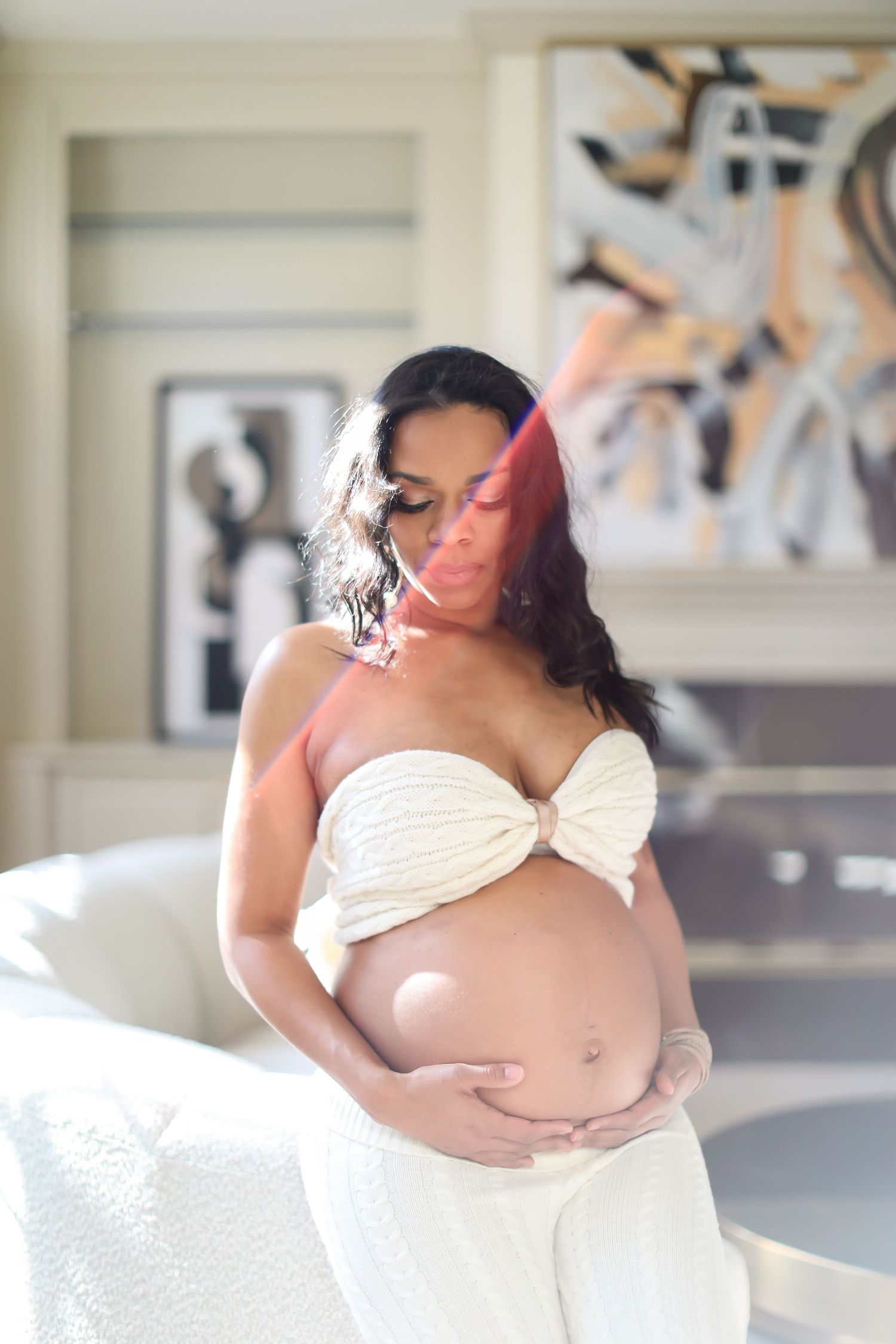 A person wearing a white outfit poses gracefully indoors for a professional maternity photoshoot.