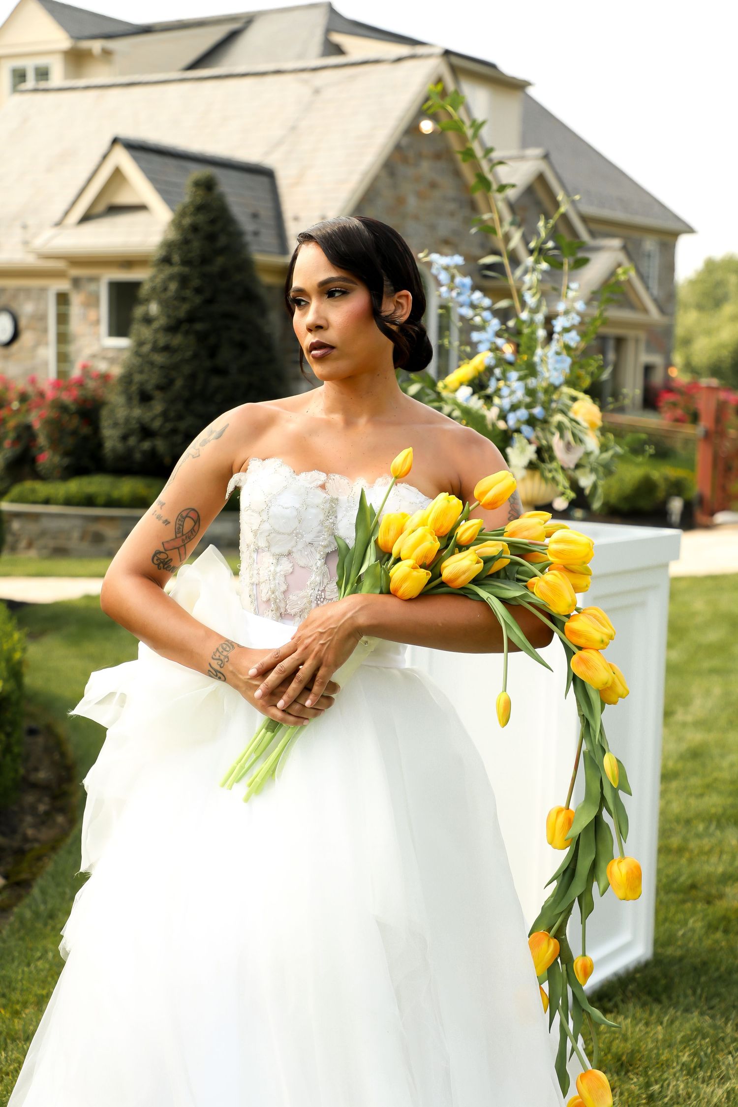A bride poses with yellow tulips in her wedding gown on a manicured lawn.