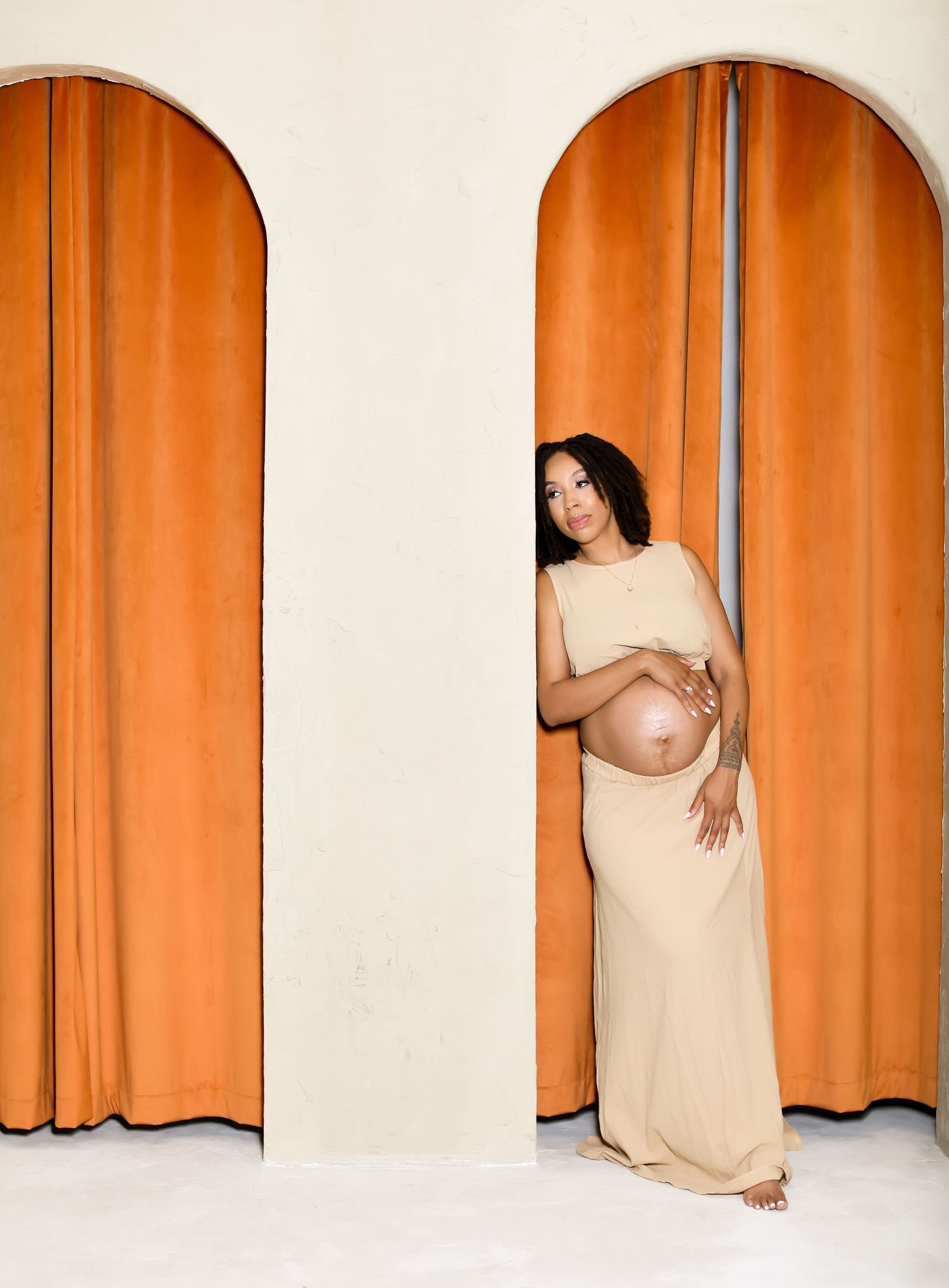 Figure in beige dress posing artistically against orange curtained archways.