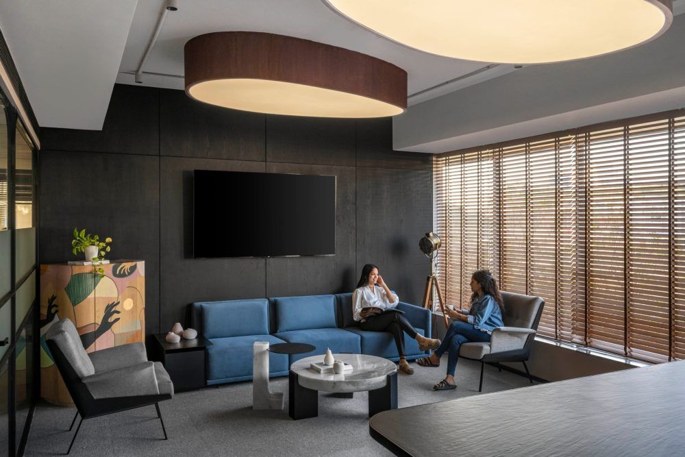 Two colleagues meeting in a stylish office lounge with blue couch, round ceiling light, and blinds.