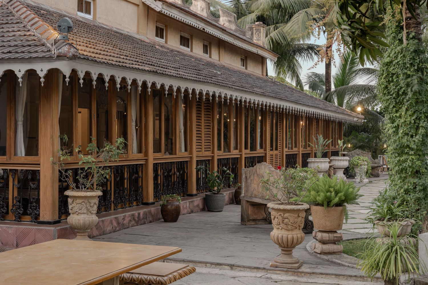 Traditional ornate wooden colonnade exterior with decorative railings, potted urns, and tropical palm trees.
