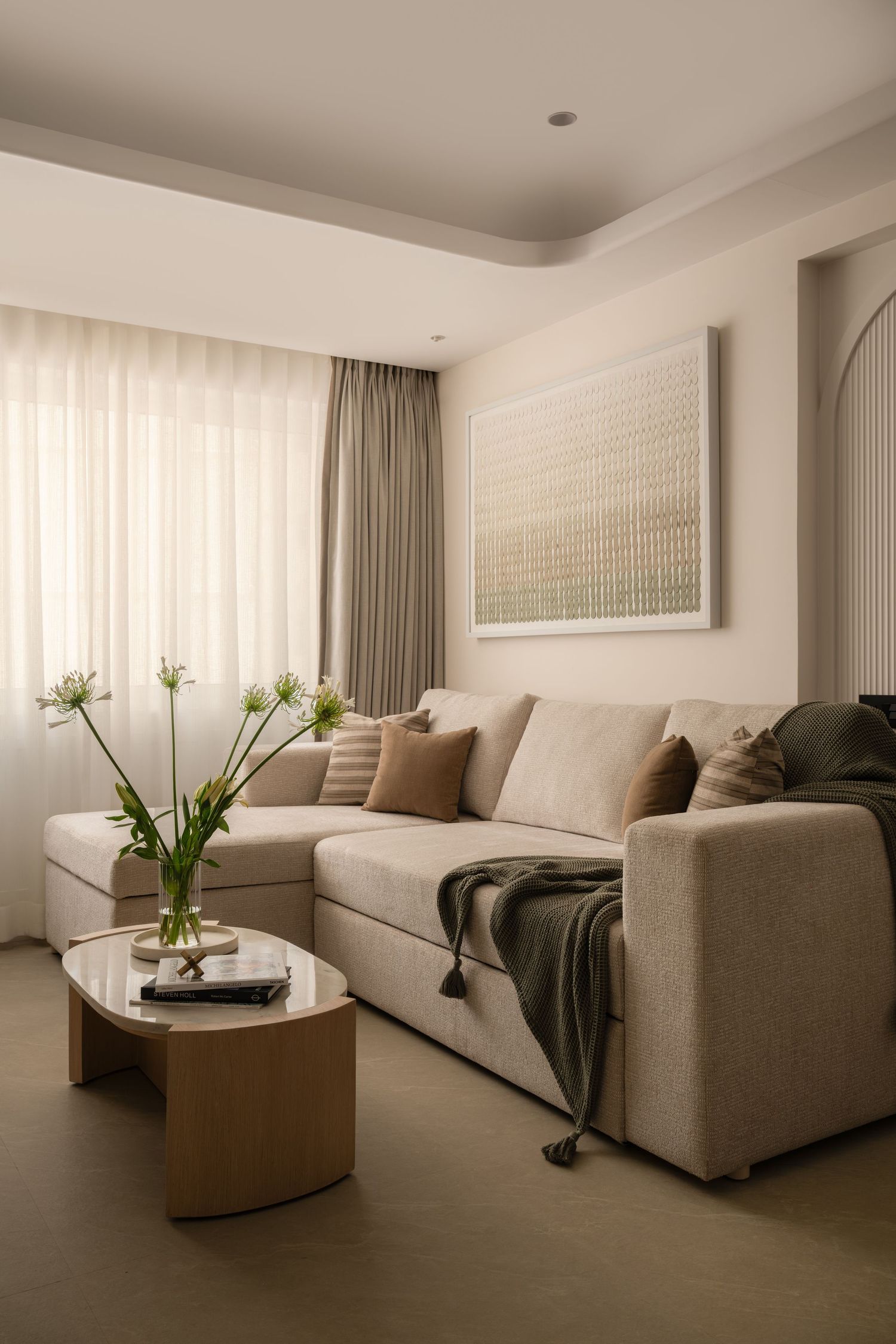 Neutral minimalist living room with beige sectional sofa, wooden coffee table, green throw, and abstract wall art.