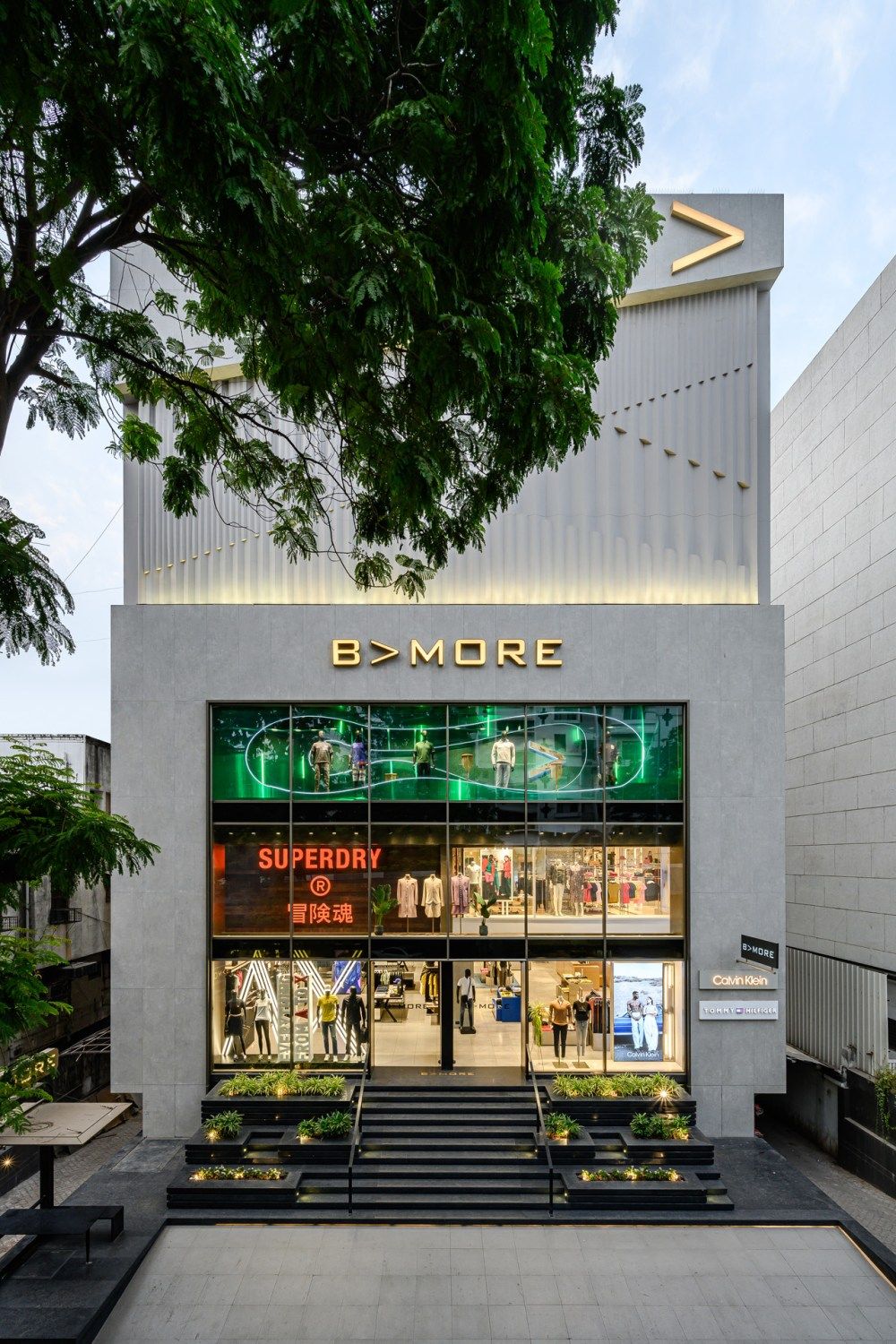 B>MORE fashion retail storefront with Superdry branding, geometric facade, and illuminated green display window.