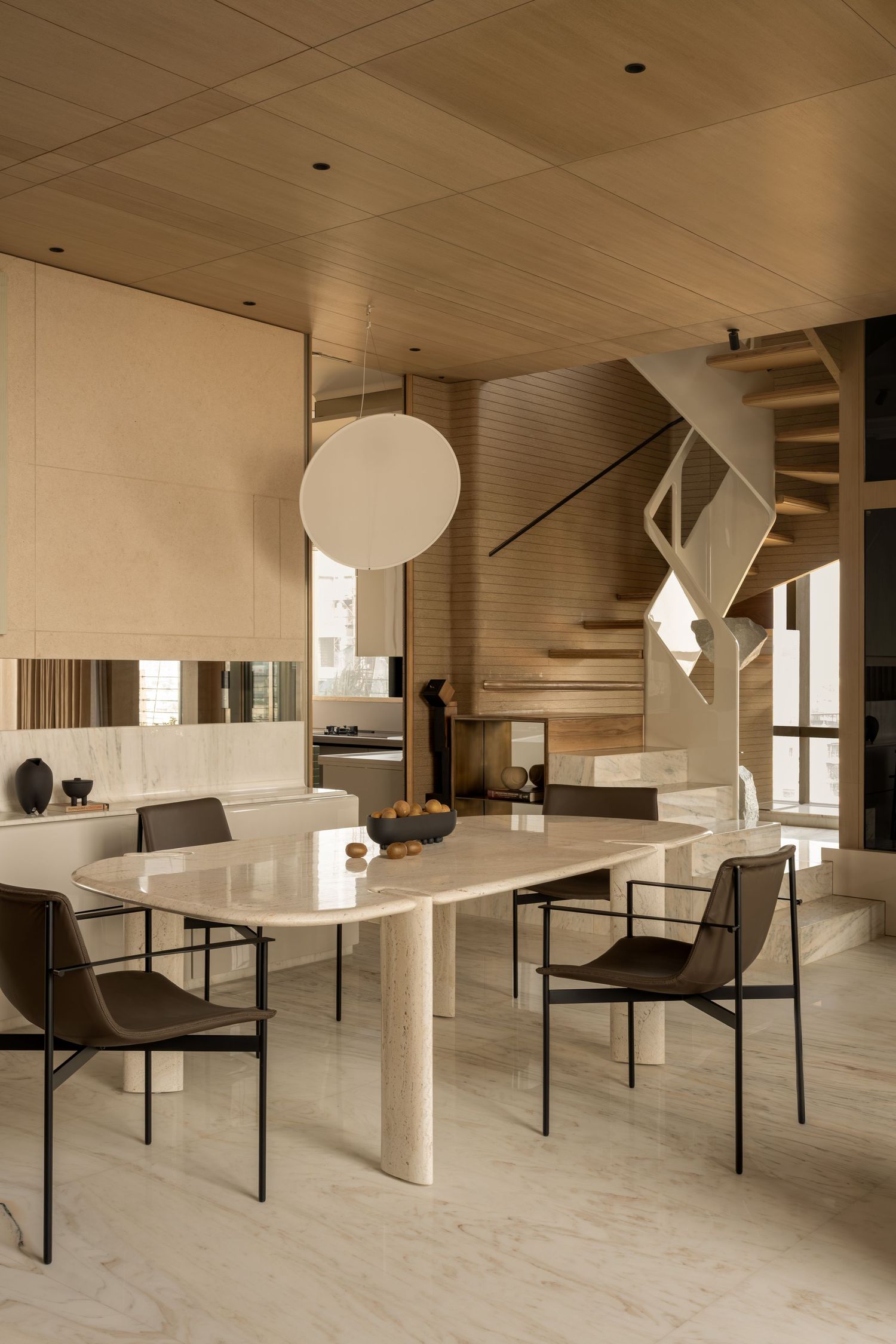 Modern dining area with travertine table, black wire chairs, globe pendant light, and sculptural open staircase.