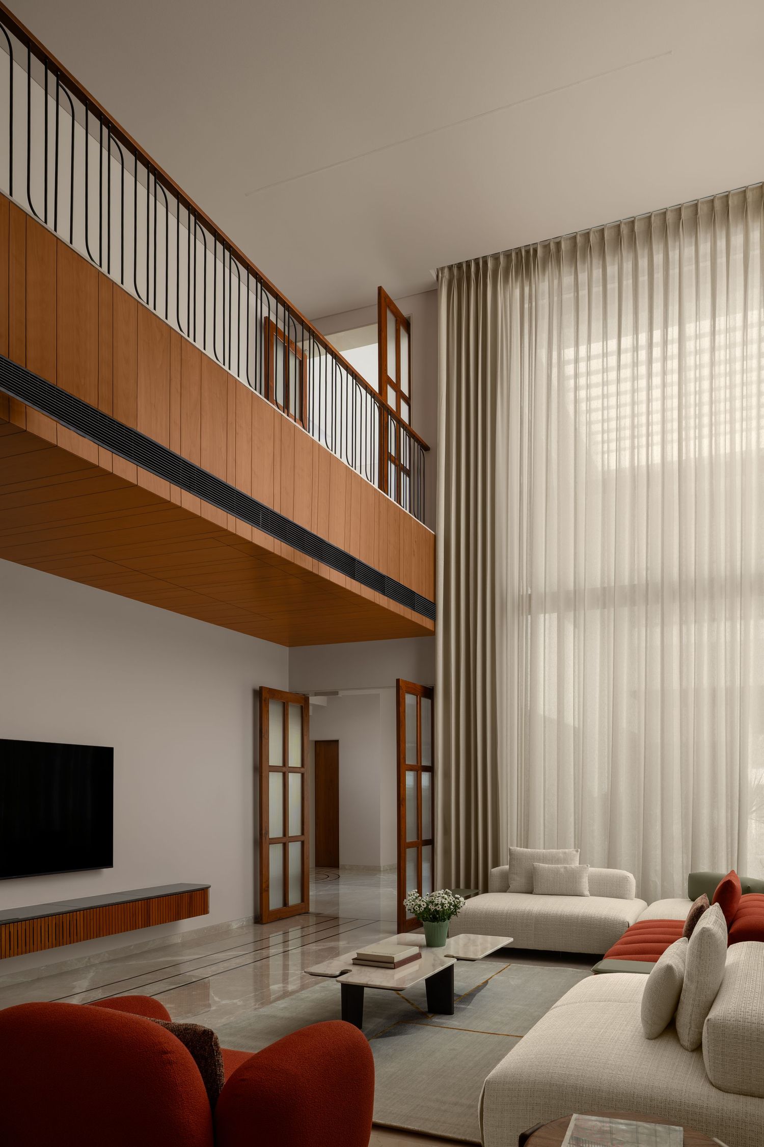 Double-height living room with mezzanine balcony, wooden railings, red accent sofa, sheer curtains, and marble floors.