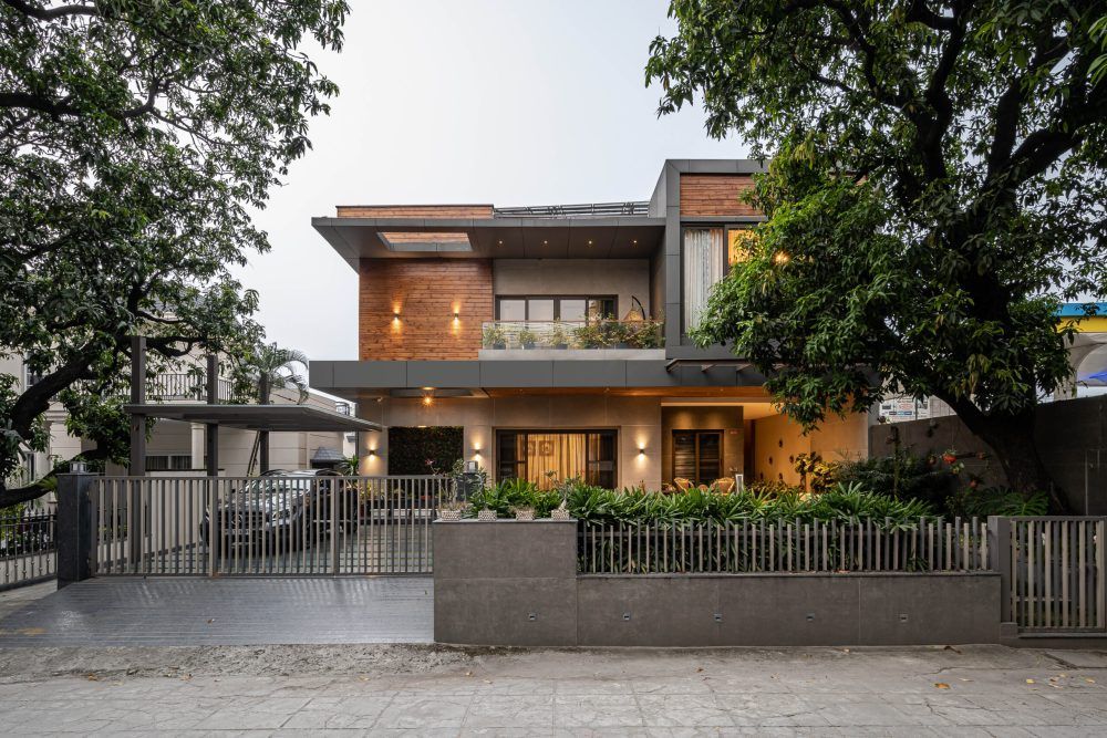 Two-story contemporary Indian home with wooden accents, balcony, mature trees, and decorative iron gate at twilight.