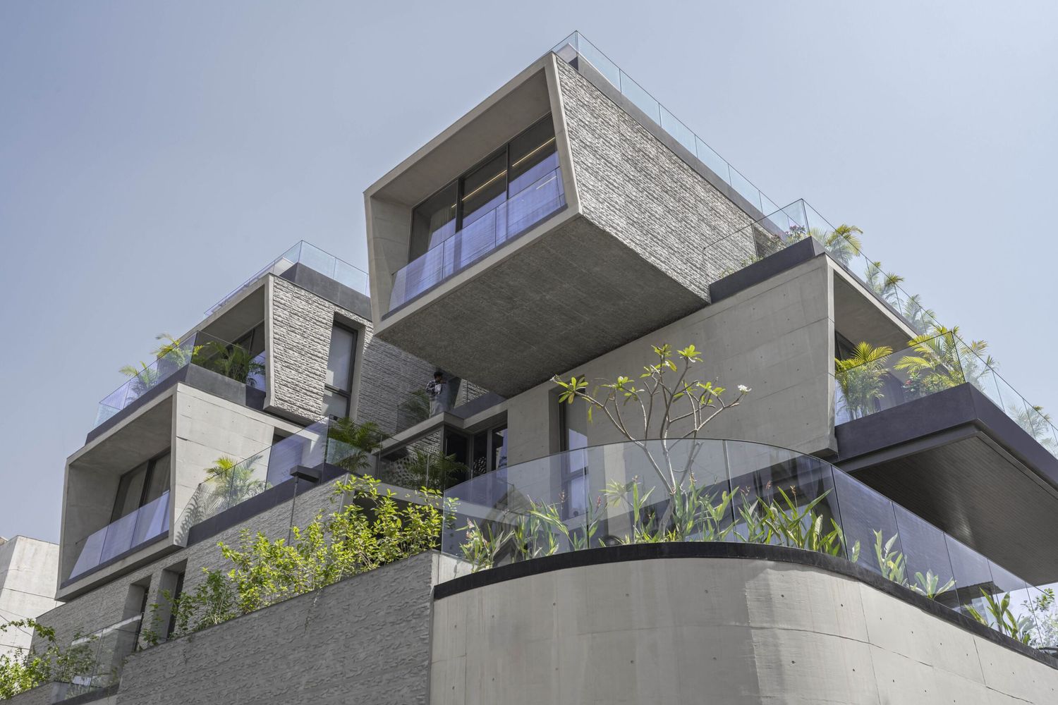 Upward perspective of modern concrete residential building featuring staggered balconies with lush green plants and glass railings.