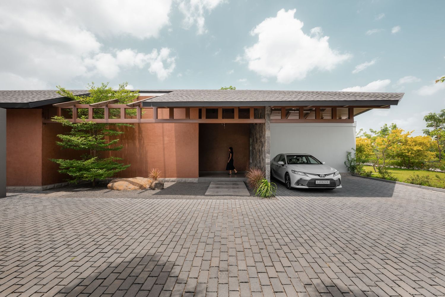 Modern single-story home with rust-colored facade, wooden pergola details, paver driveway, and white car parked under covered entry.