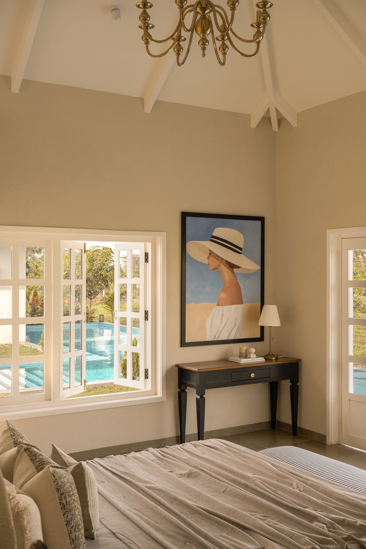 Serene bedroom with vaulted ceiling, portrait artwork, pool view window, brass chandelier, and neutral bedding.