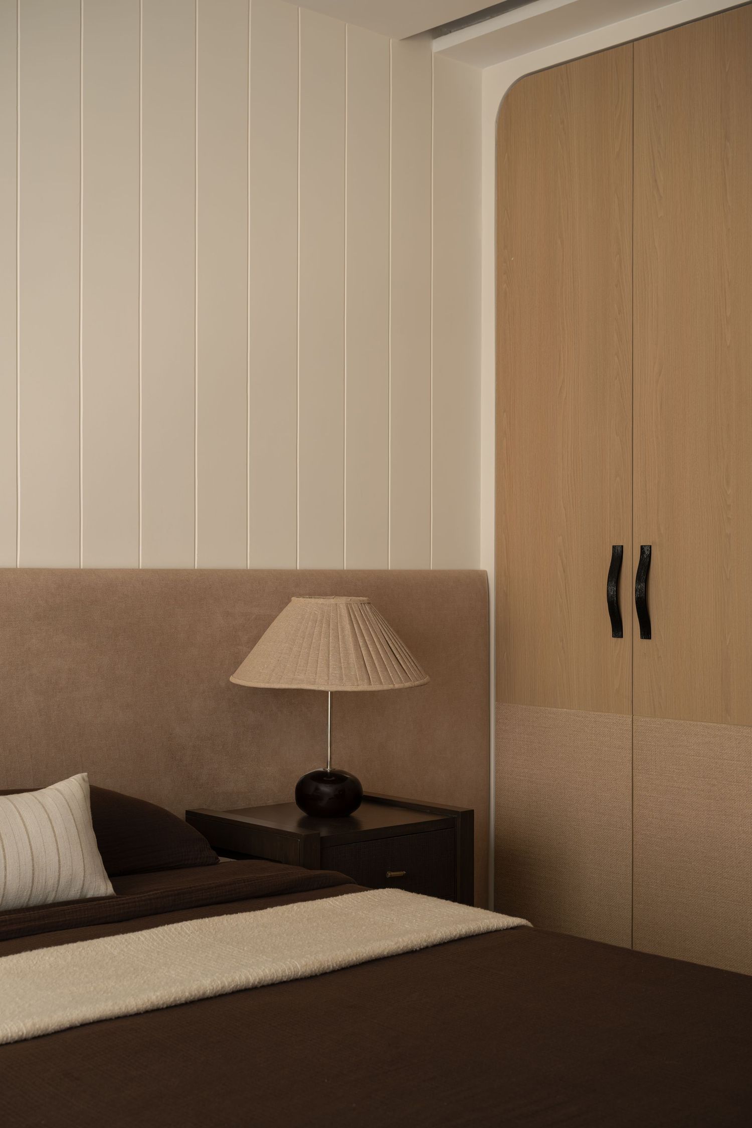 Modern bedroom with wood wardrobe, pleated lamp, dark headboard, and vertical white paneled wall.
