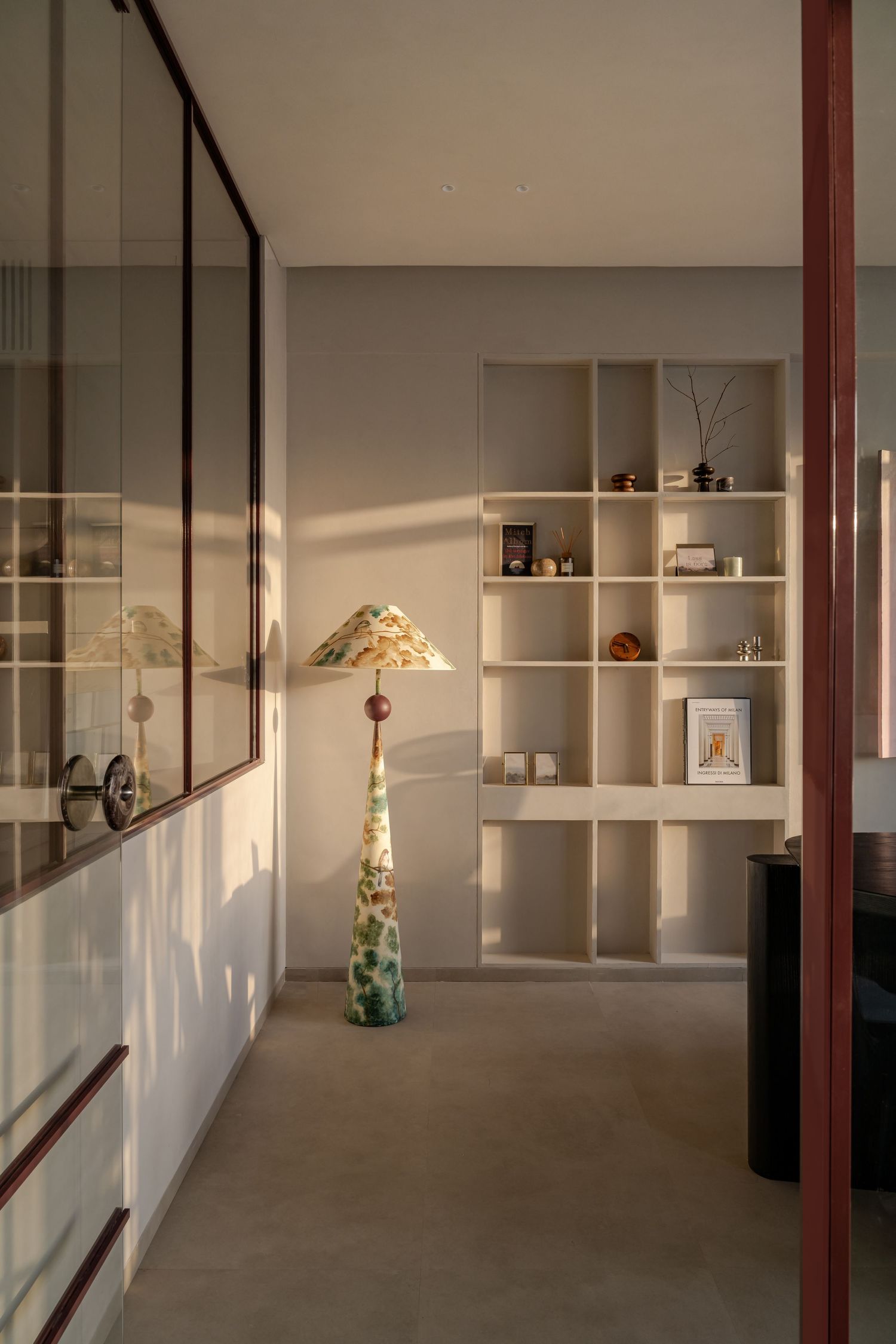 Contemporary hallway with glass partition, decorative floor lamp, and built-in cream shelving unit with curated objects.