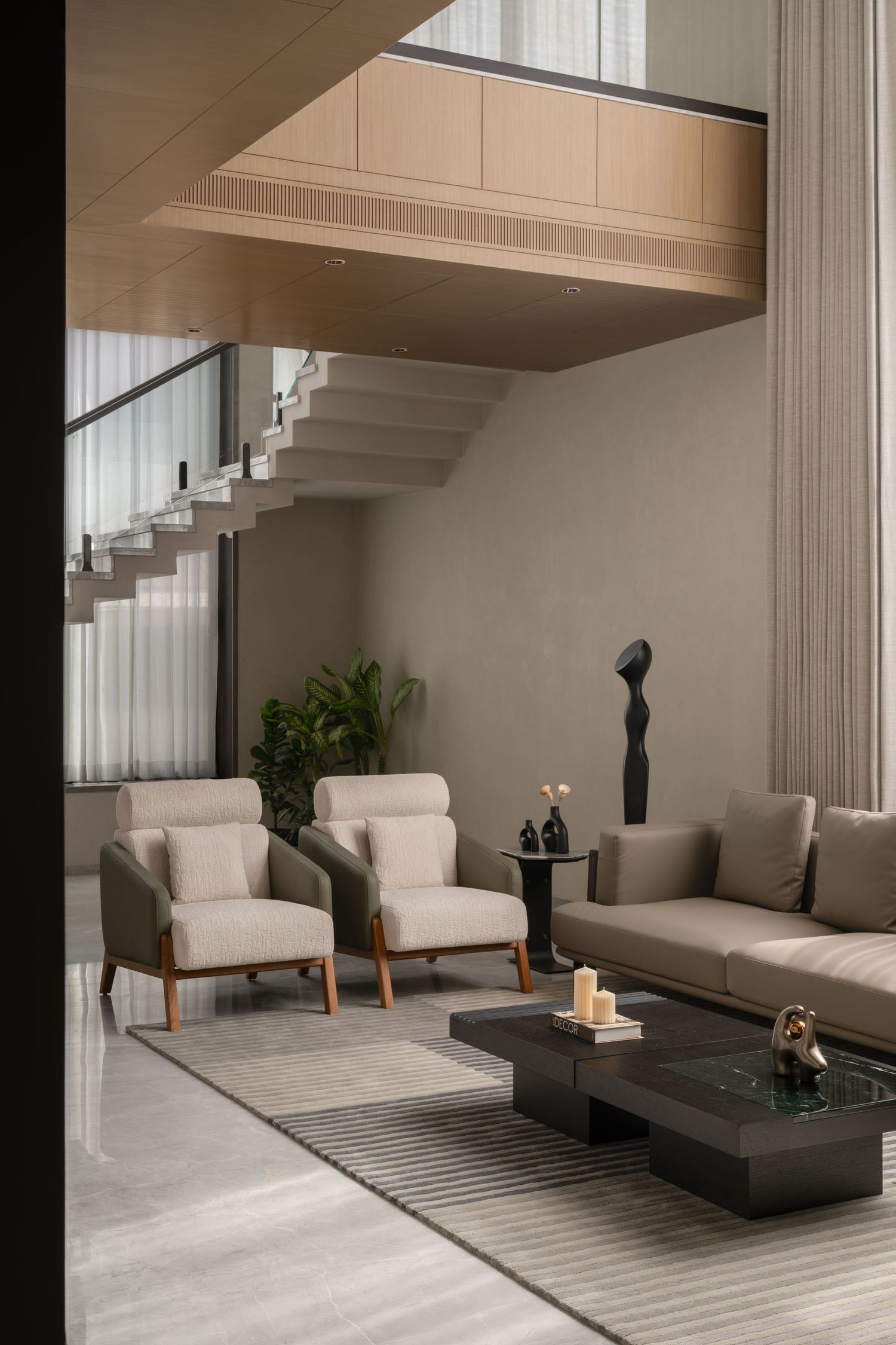 Modern minimalist living room with beige sofas, wood armchairs, dark coffee table, floating staircase, and sculptural decor.