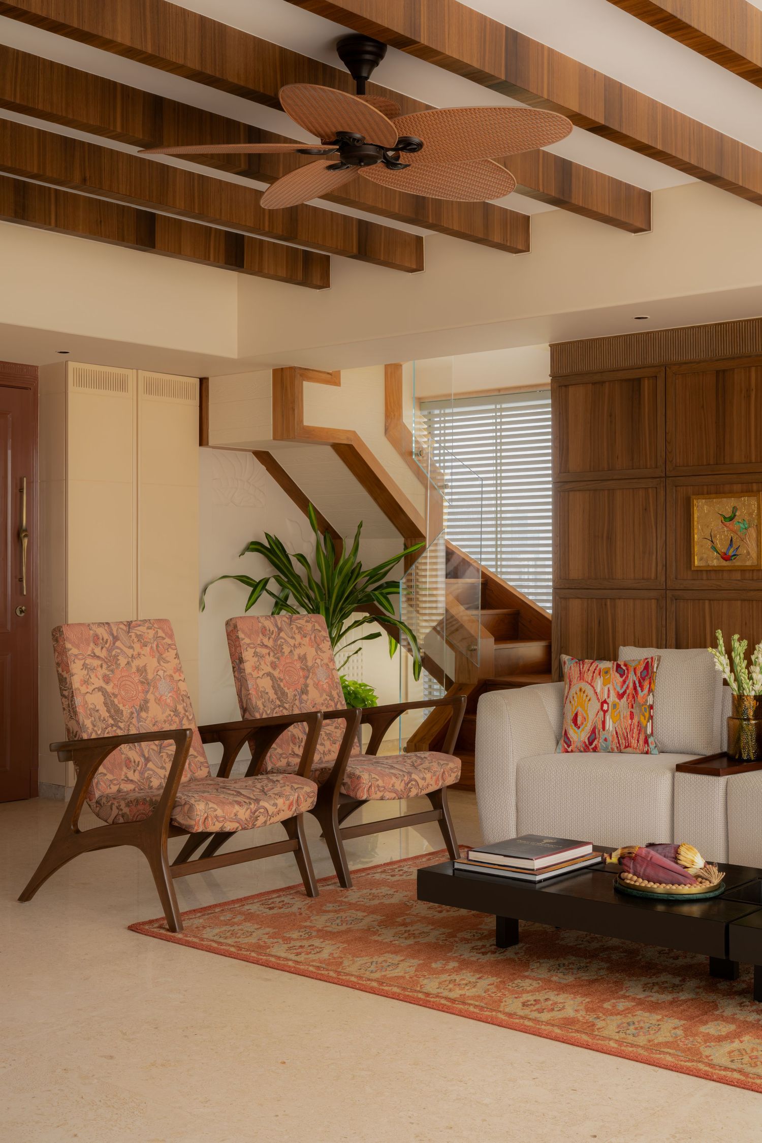 Warm living room with wooden beam ceiling, patterned chairs, ceiling fan, and staircase.