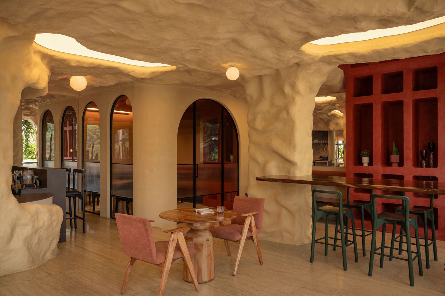 Cave-like restaurant interior with arched doorways, textured cream walls, pink chairs, and a red bar area.