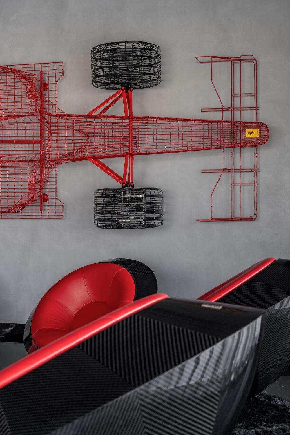 Close-up of a red wire Ferrari F1 car sculpture mounted on a gray concrete wall above red chairs.
