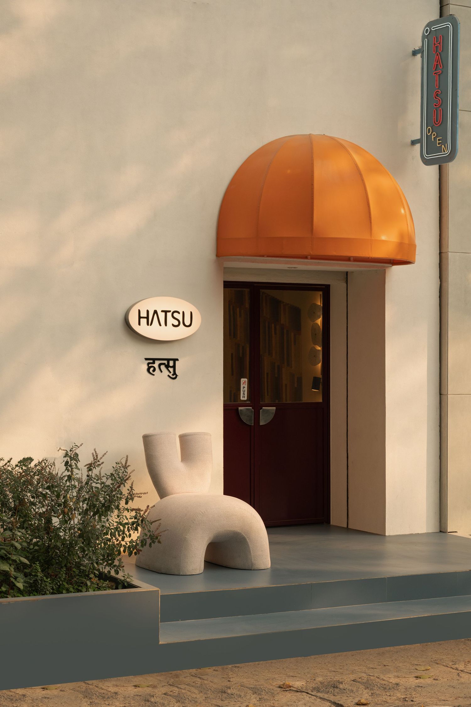 Modern restaurant entrance named Hatsu with orange dome awning, sculptural white chair, and Hindi signage.