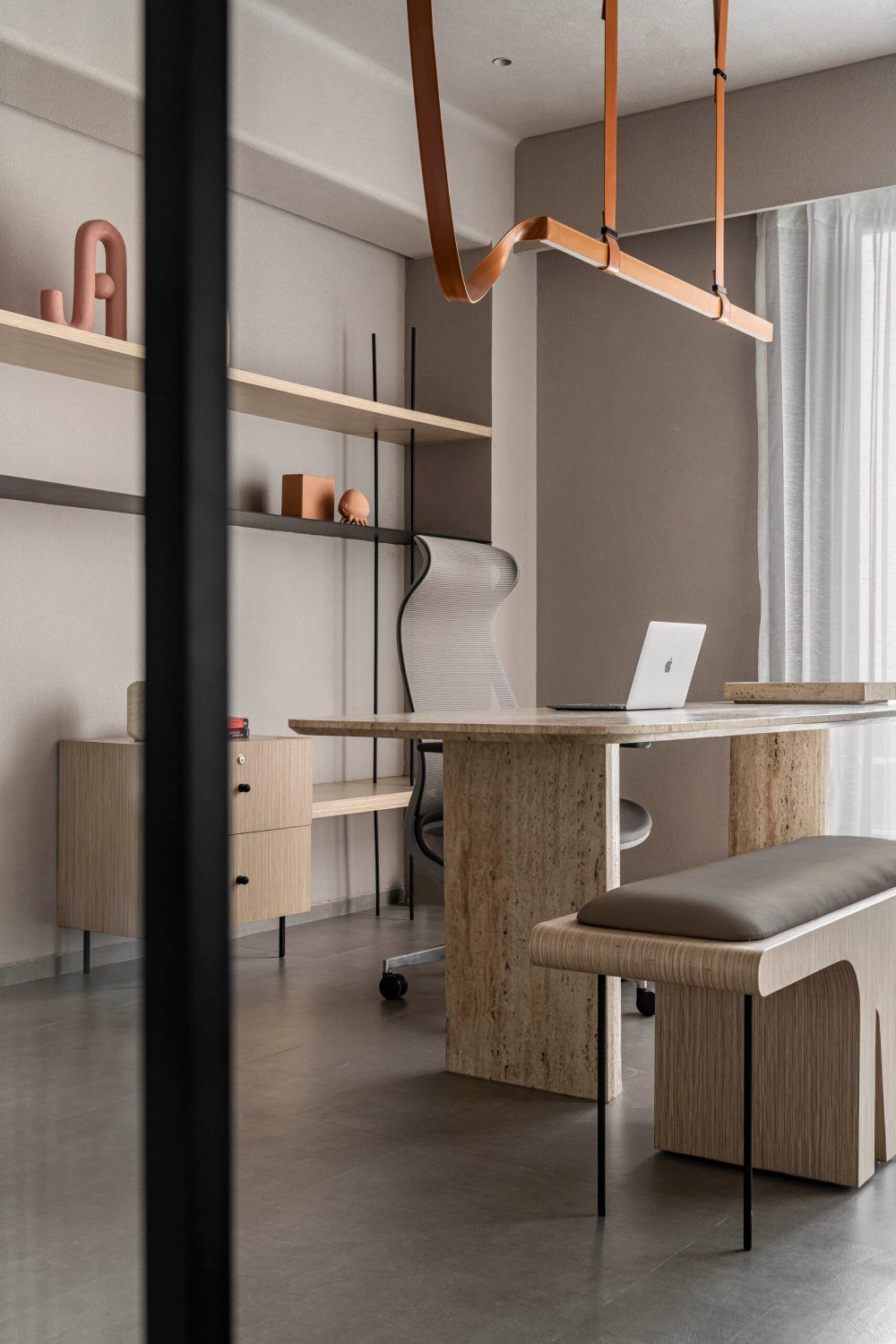 Minimalist office workspace with travertine desk, leather pendant light, and open shelving unit.