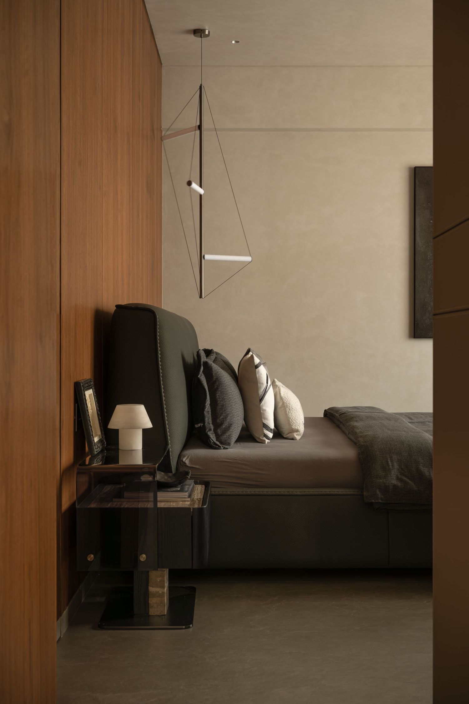 Minimalist bedroom viewed through doorway with dark upholstered bed, geometric pendant light, and warm wood panel wall.