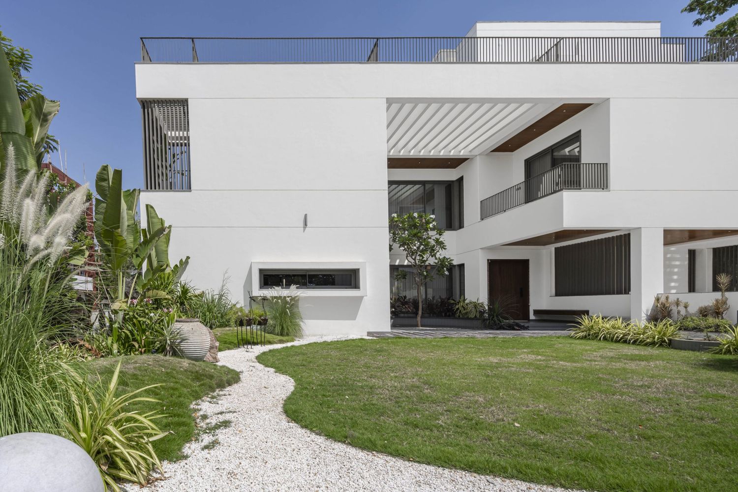 Modern white villa with clean lines, lush garden, gravel walkway, and spacious balcony under a blue sky.
