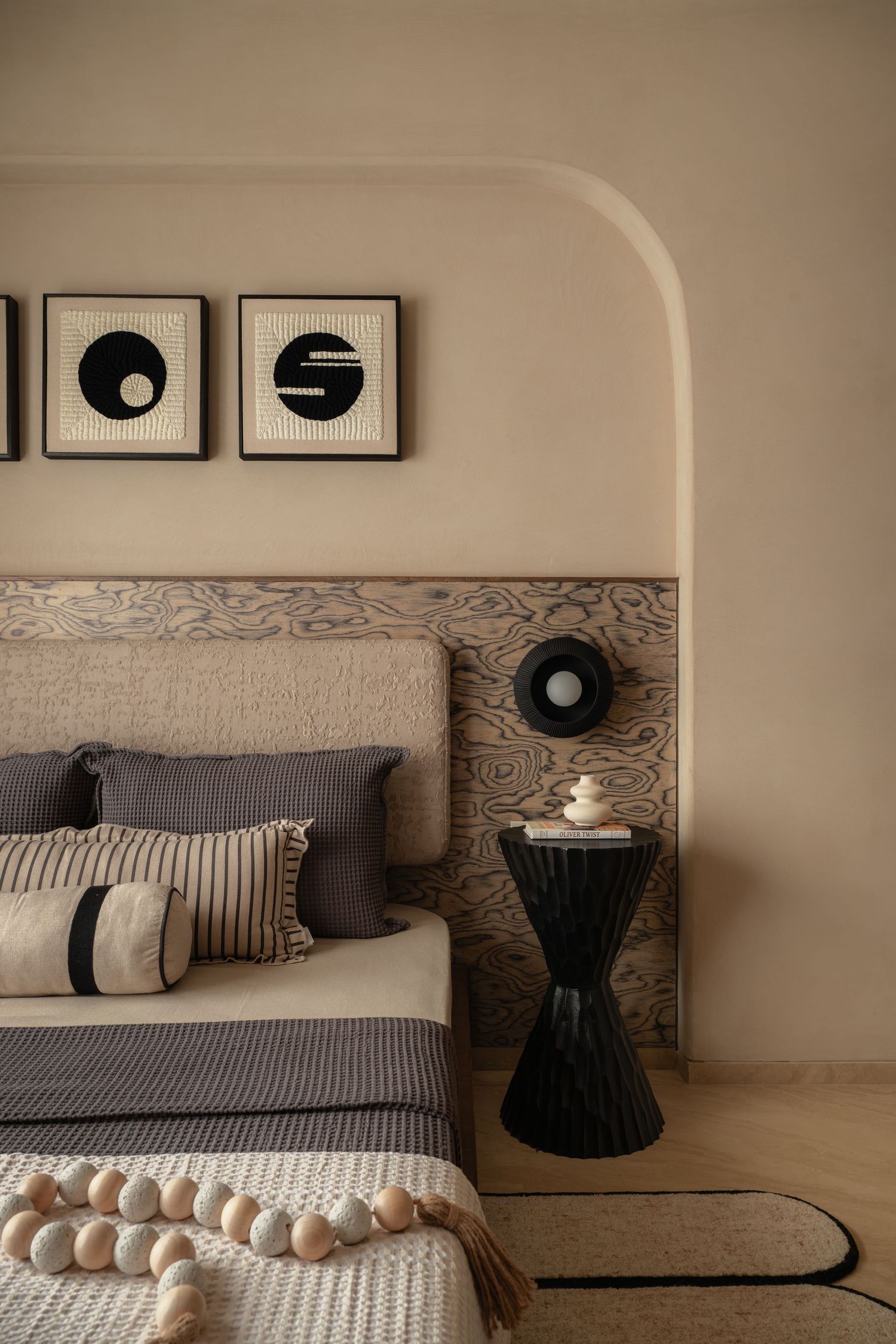 Minimalist bedroom with wooden headboard, striped pillows, black side table, and abstract wall art.