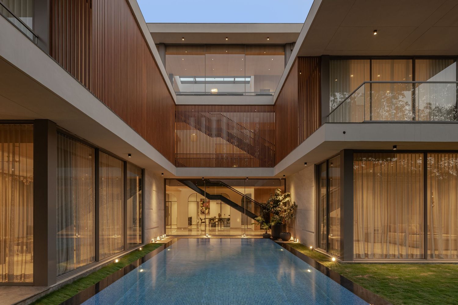 Luxury modern villa exterior at dusk featuring wood cladding, glass walls, illuminated lap pool, and open courtyard.