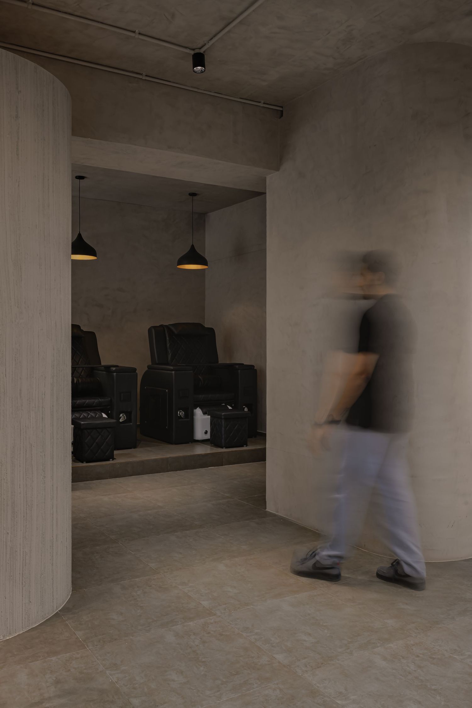 Moody salon corridor with curved wooden wall panel, blurred figure walking past a room with black reclining chairs and pendant lights.