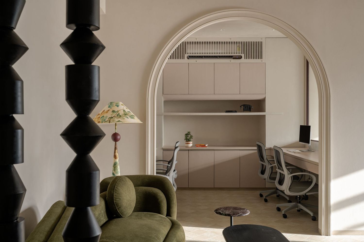 Modern home office with arched doorway, beige cabinetry, ergonomic chairs, green velvet sofa, and decorative floral lamp.