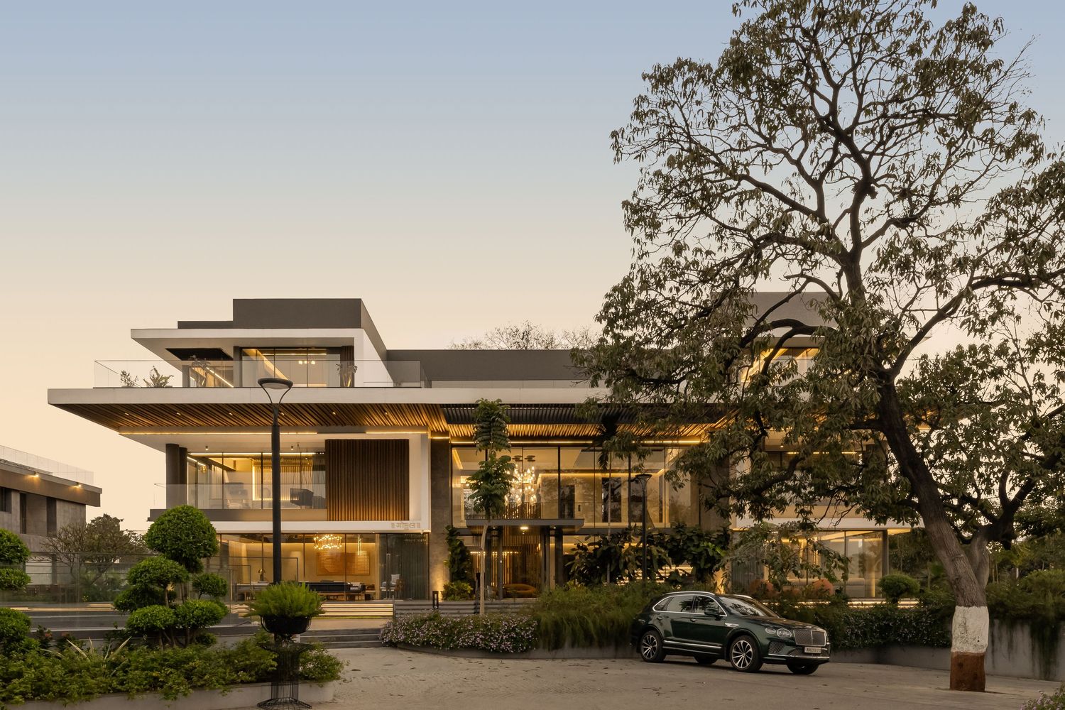 Modern luxury villa with glass facades, flat roof, lush trees, and a parked car at dusk in a landscaped setting.