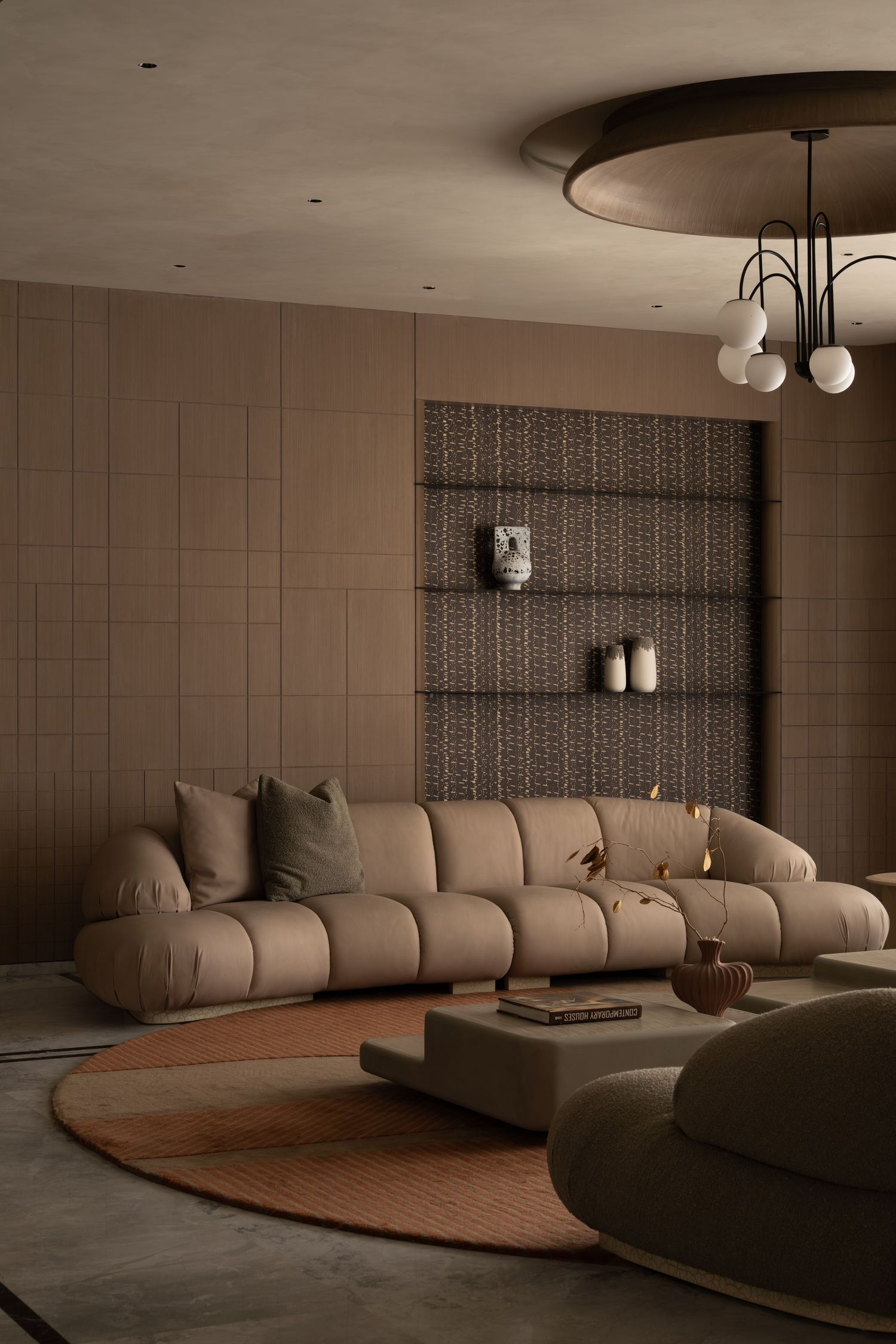 Luxurious curved beige sofa on round rug beneath ornate ceiling fixture with globe lights and wall shelving.