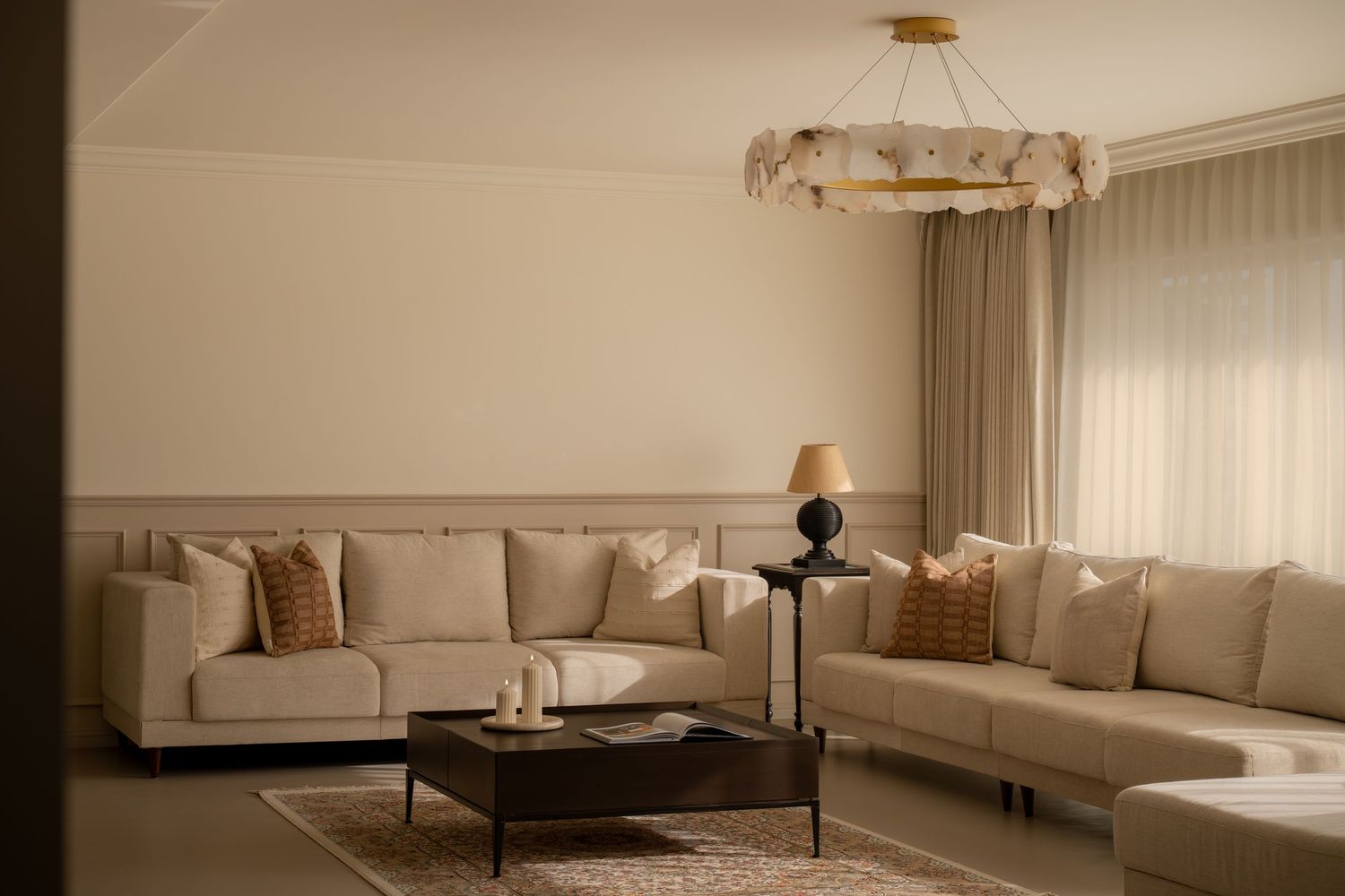 Elegant living room with beige sofas, gold chandelier, amber accent pillows, dark coffee table, and sheer curtains.