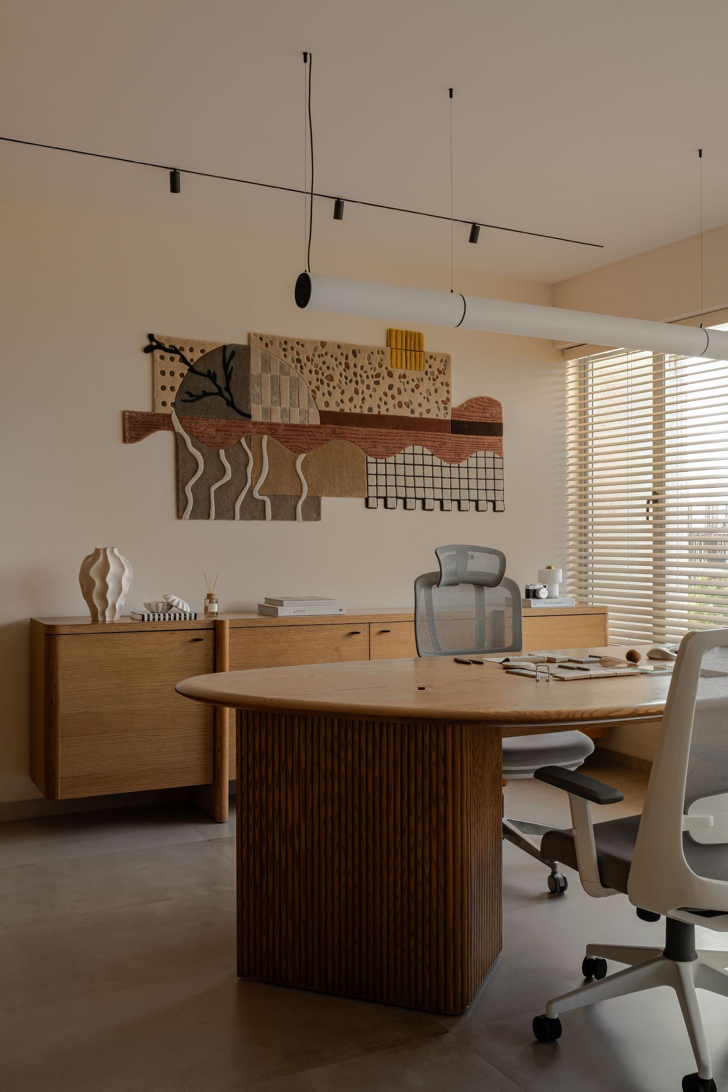 Minimalist office with oval fluted wood desk, ergonomic chairs, abstract wall art, and warm beige tones with blinds.