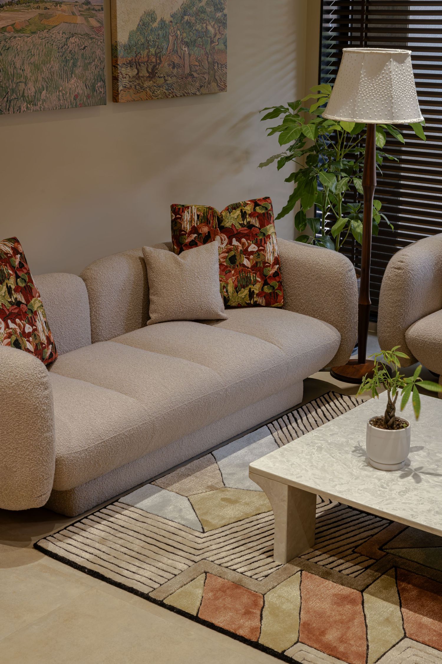 Cozy living room with a cream boucle sofa, floral pillows, marble coffee table, floor lamp, and lush green plant on a patterned rug.