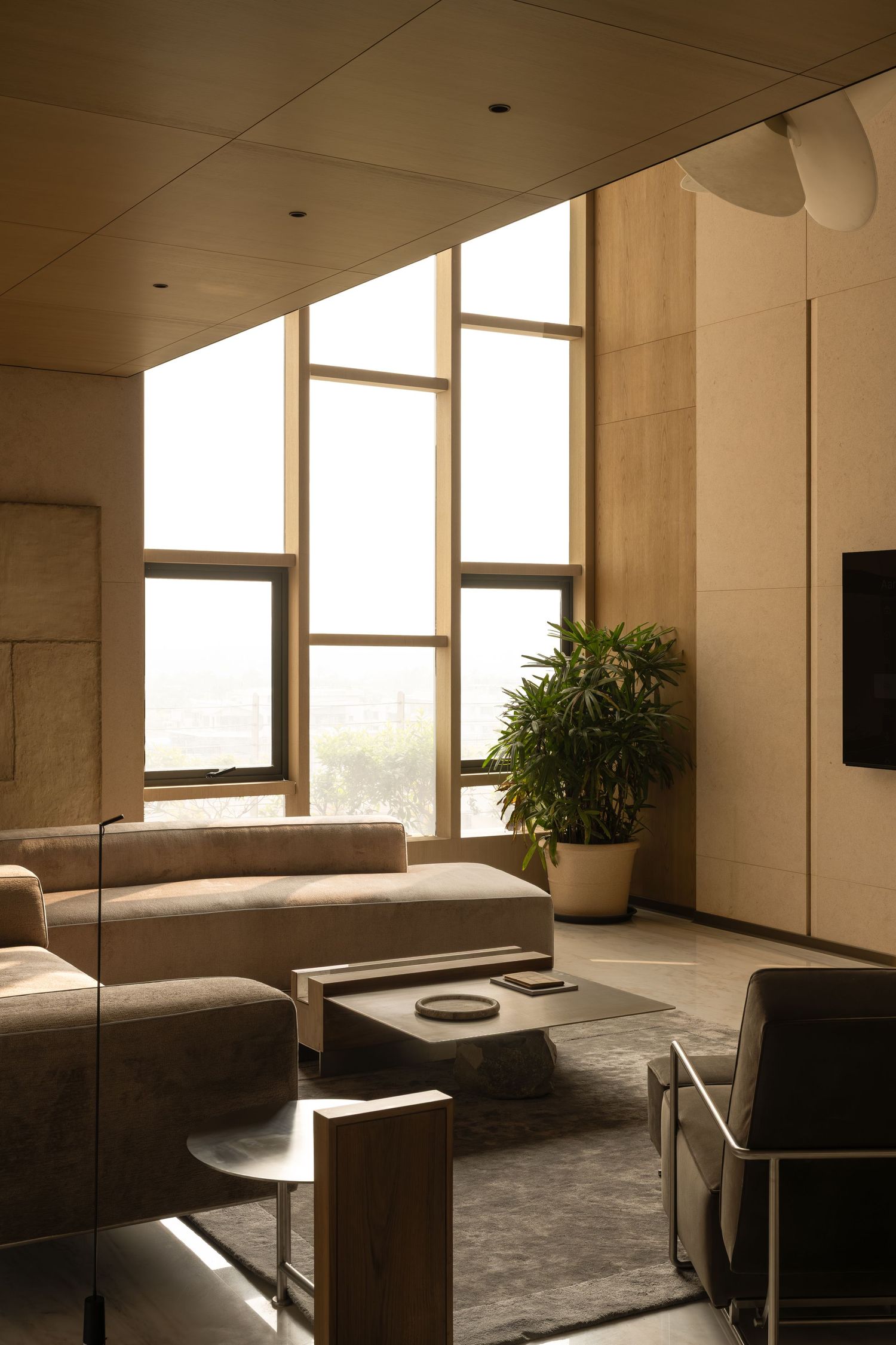 Spacious sunlit living room with beige sofas, large floor-to-ceiling windows, tropical plant, and dark area rug.