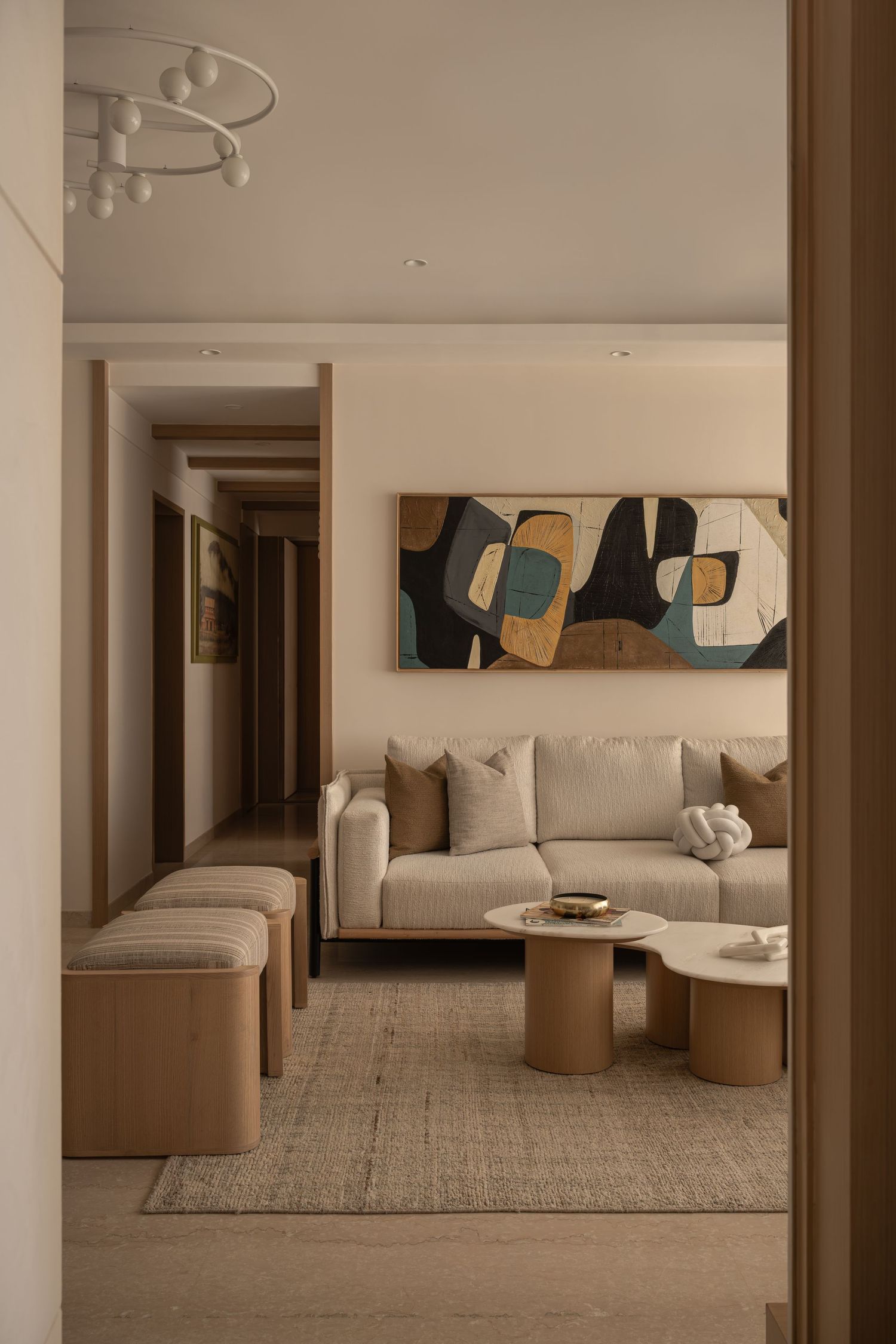 Contemporary living room with beige sofa, oval coffee tables, abstract artwork, and wooden ottomans.