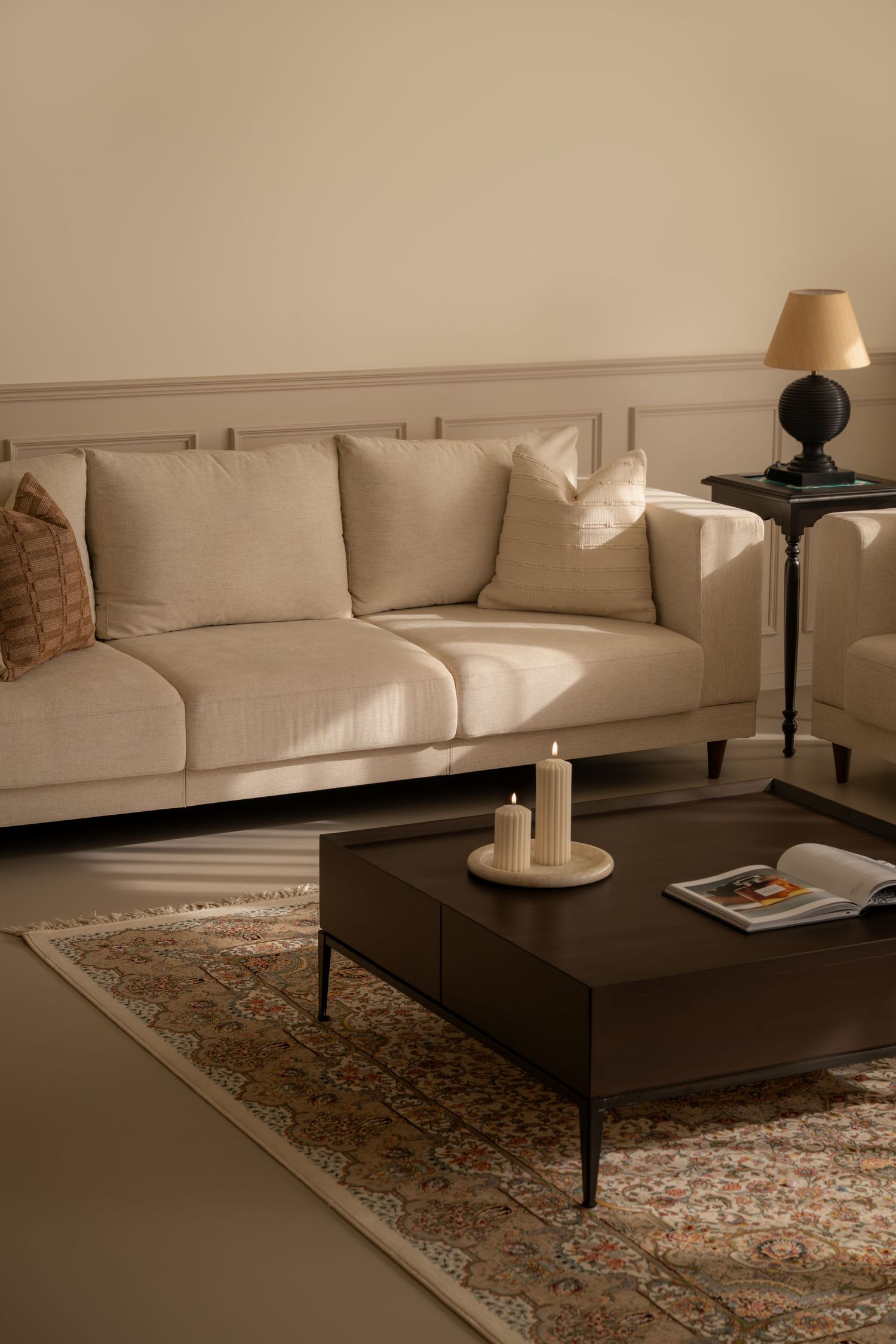 Elegant neutral living room with beige linen sofa, dark coffee table, candles, patterned rug, and black table lamp.