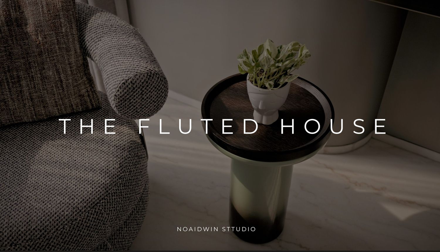 A dark modern side table with a small plant beside a gray sofa, featuring The Fluted House branding by Noaidwin Studio.