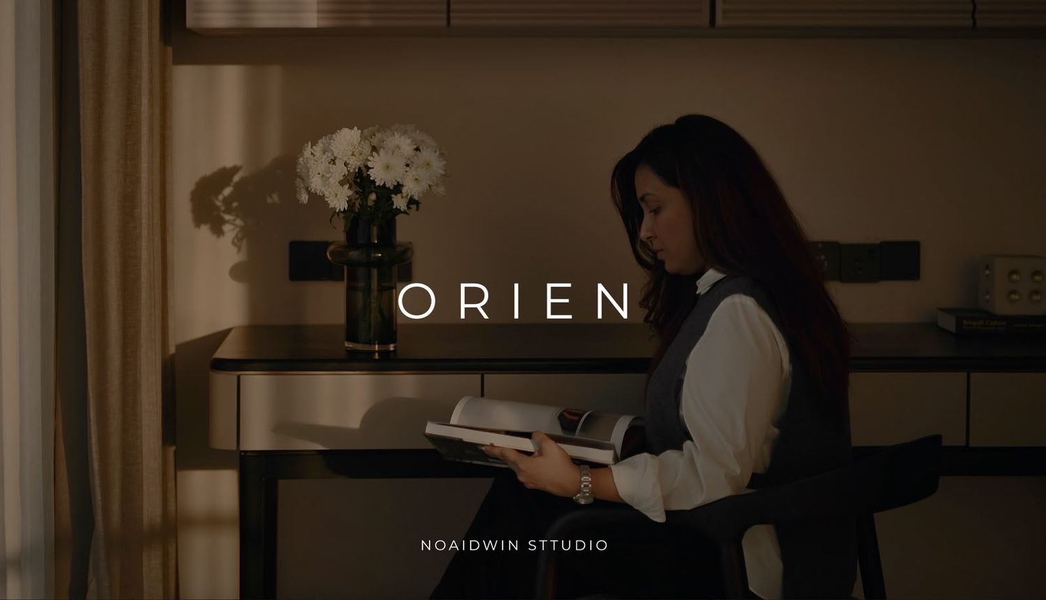 A woman reads a book in a warmly lit room beside a desk with flowers, overlaid with the text ORIEN.