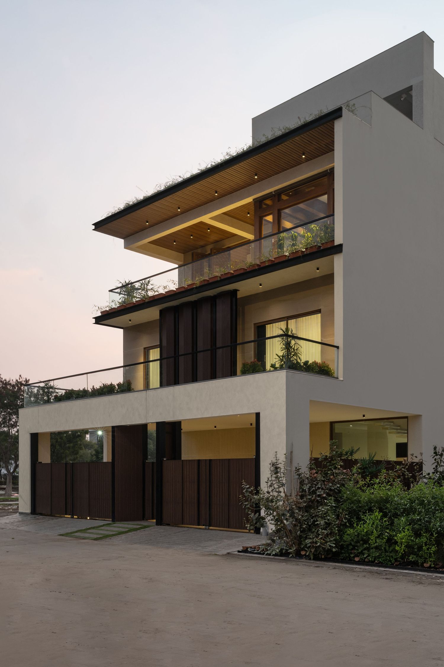 Contemporary three-story home with white render, dark wood screens, cantilevered roof, and rooftop garden at dusk.