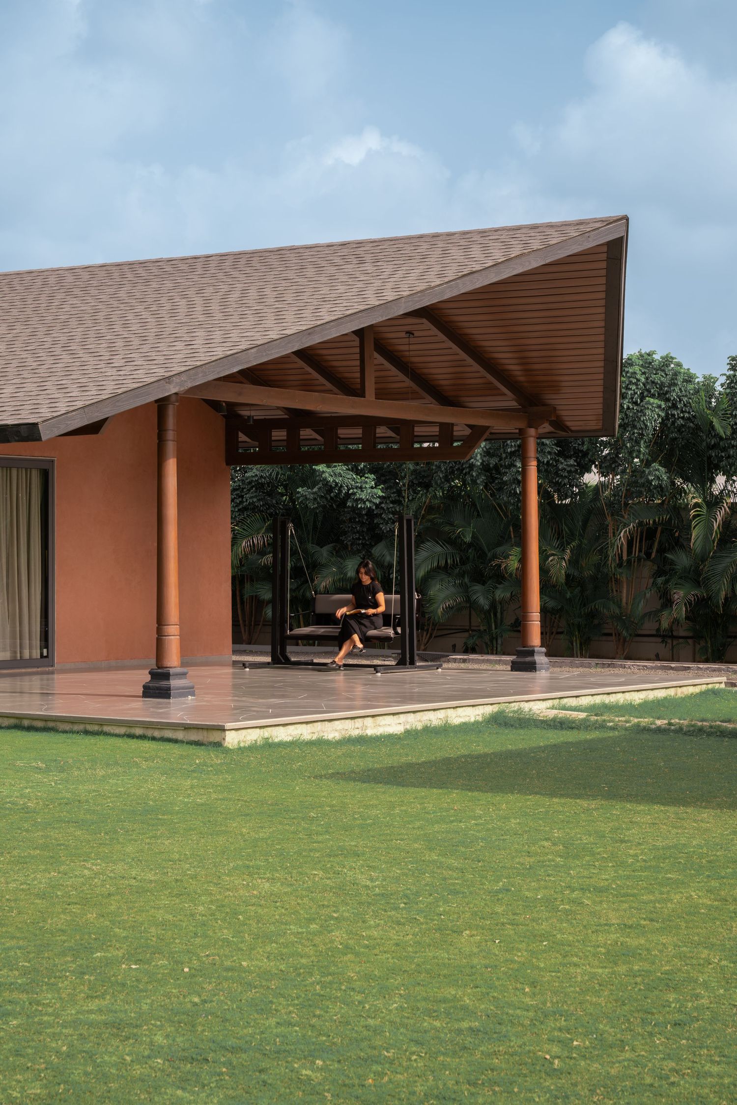 Terracotta pavilion with pitched roof, wooden columns, swing seat, and lush green lawn surrounded by tropical trees.