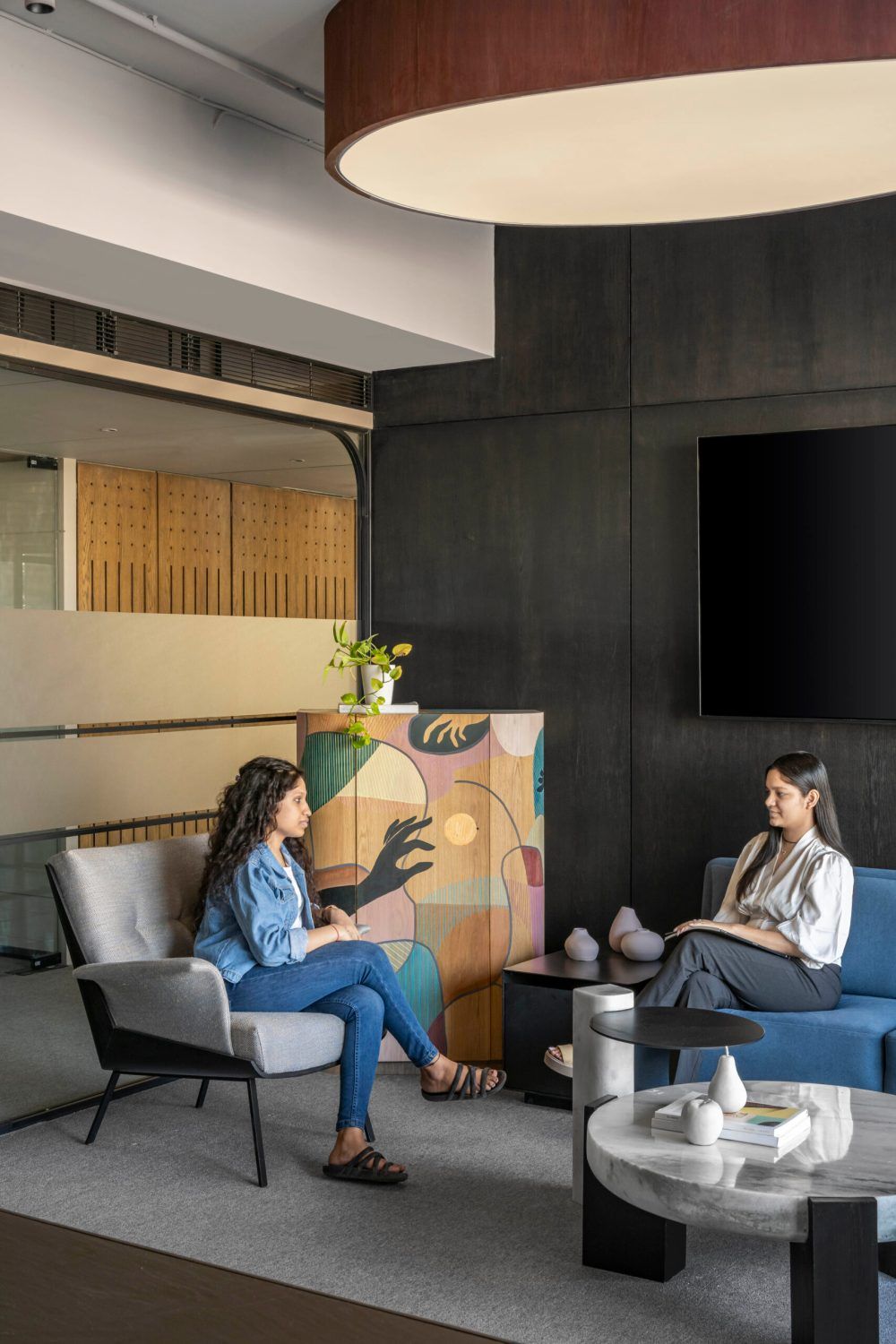 Two women having a conversation in a contemporary office lounge with blue sofa and artistic decor.
